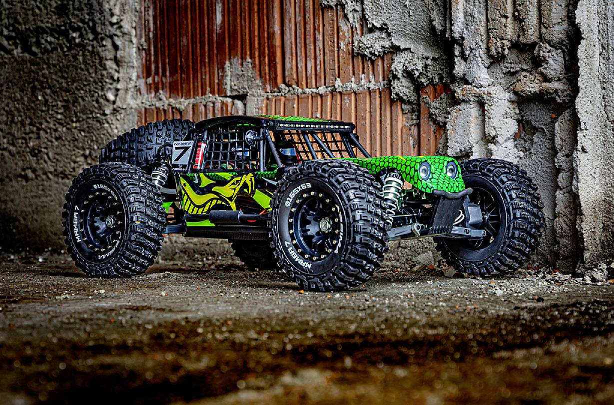 Une voiture télécommandée avec de grandes roues et une peinture verte en motif de serpent est placée devant un mur de béton brut dans un environnement urbain.