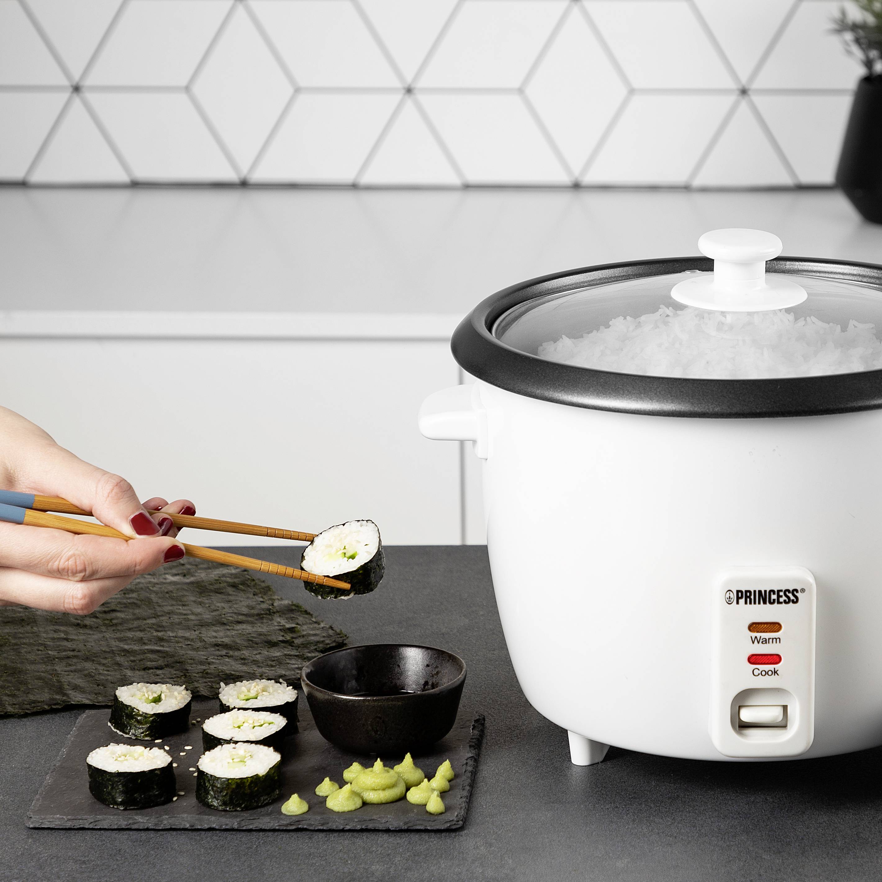 Une personne tient des rouleaux de sushi avec des baguettes au-dessus d'une table sur laquelle se trouvent un cuiseur à riz, du wasabi et de la sauce soja. En arrière-plan, des carreaux blancs.