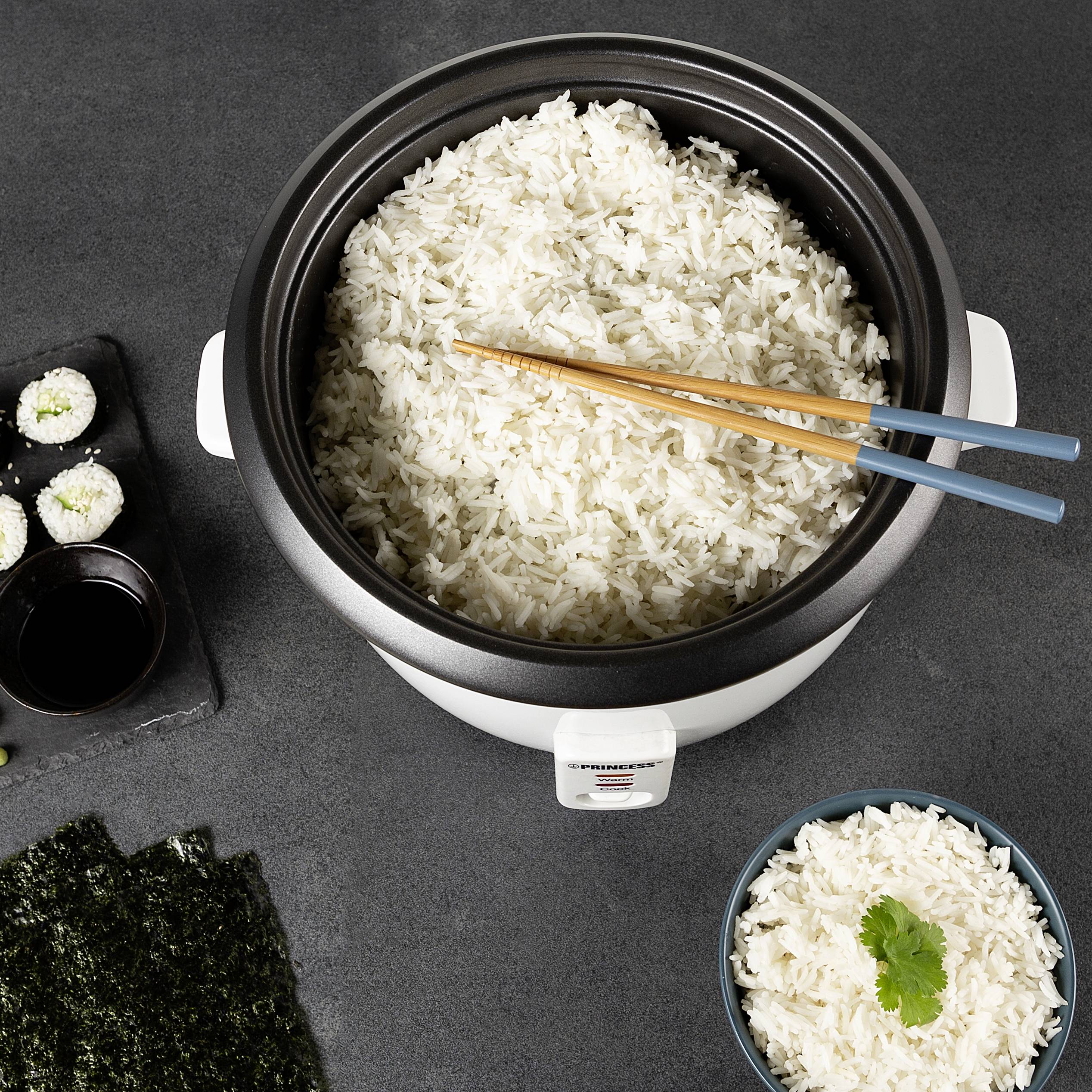 Cuiseur de riz avec du riz fraîchement cuit et des baguettes, entouré d'ingrédients à sushi et d'un petit bol de riz avec de la coriandre.