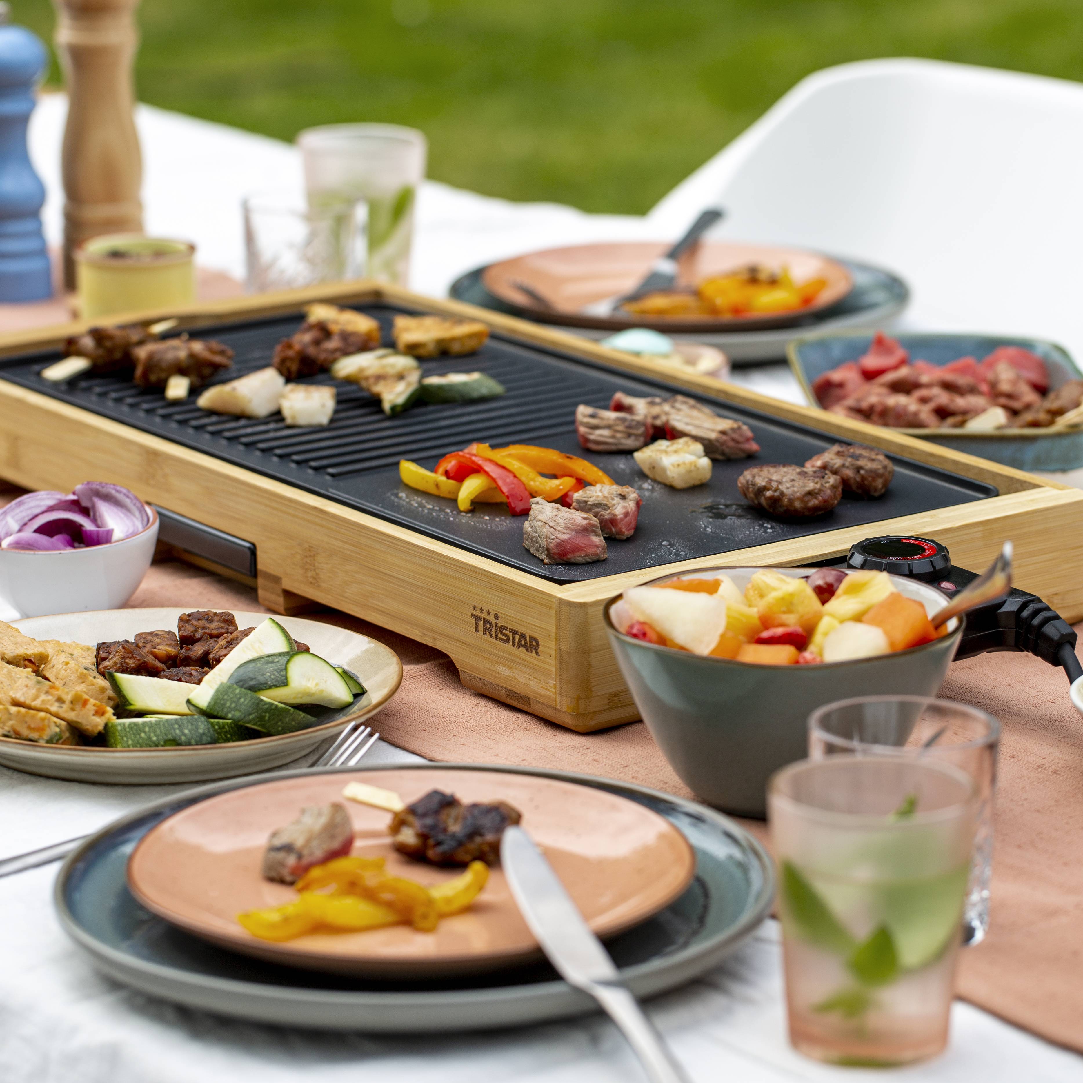Une table à l'extérieur avec une plaque de grill sur laquelle de la viande et des légumes sont en train d'être grillés. Des assiettes avec de la nourriture et des boissons sont disposées.