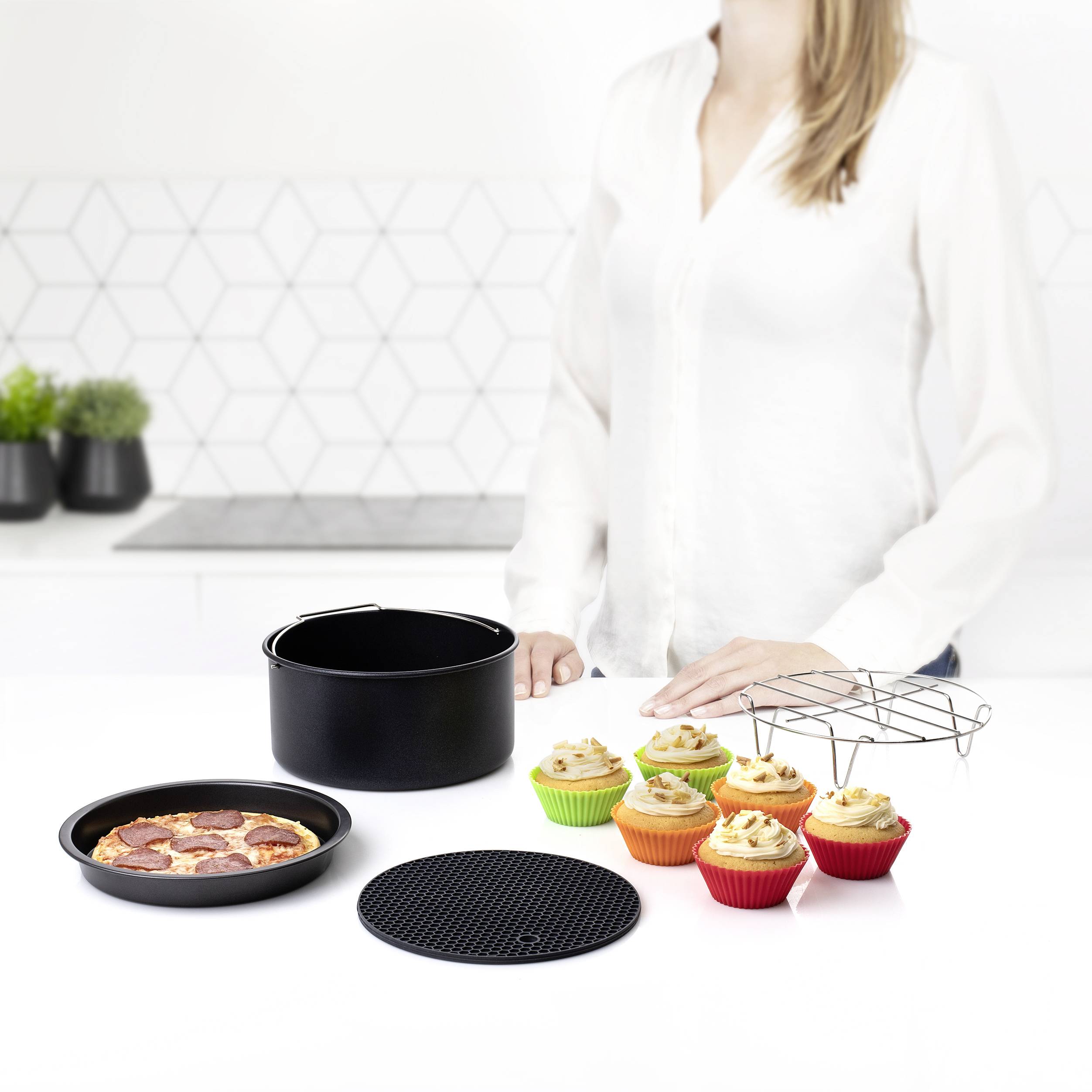 Une personne portant un haut blanc se tient près d'une table avec une pizza, des muffins colorés, une poêle noire et des dessous de plat.