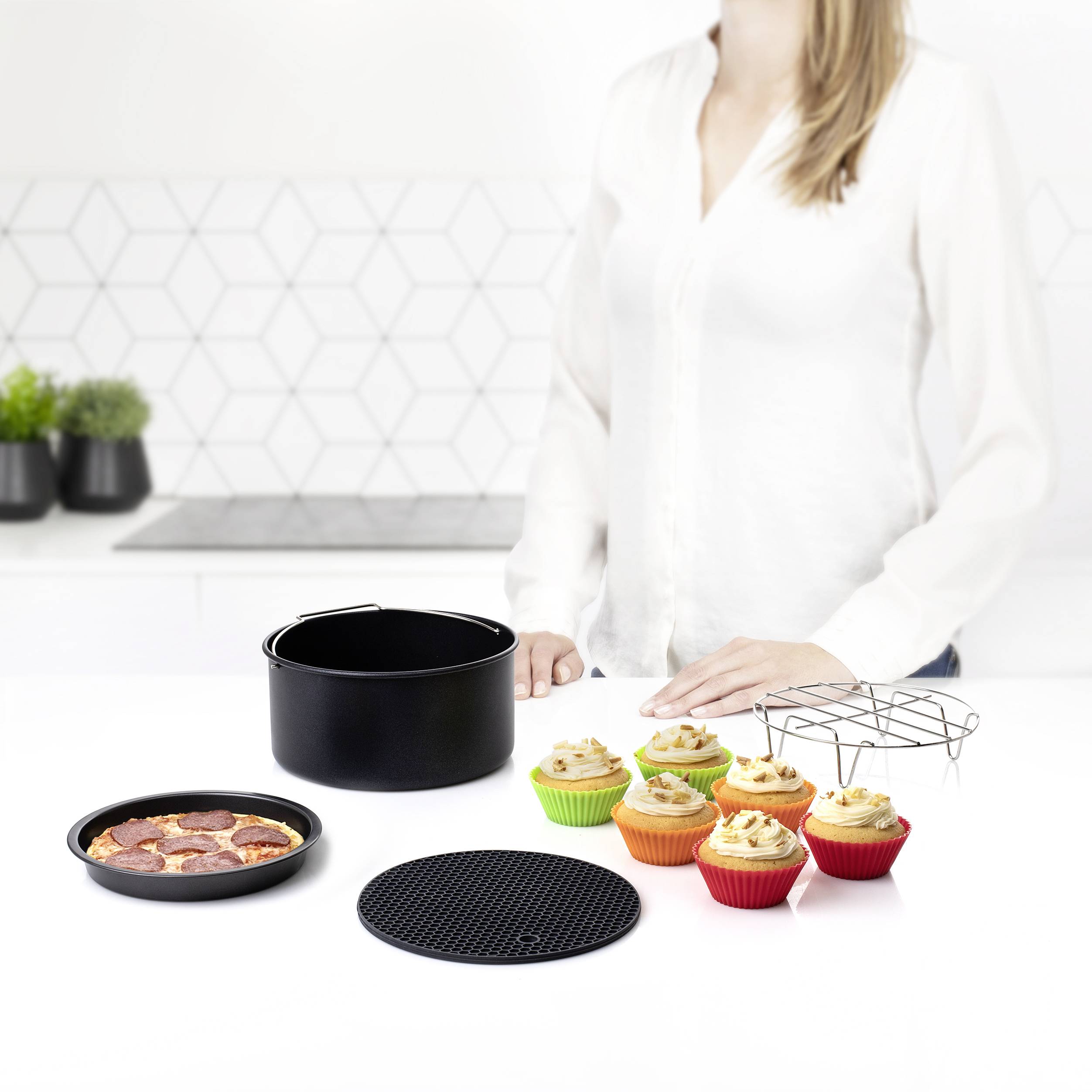 Une personne se tient dans une cuisine moderne. Sur le plan de travail, on peut voir un gâteau, des cupcakes dans des moules colorés, une grille et un moule à gâteau.