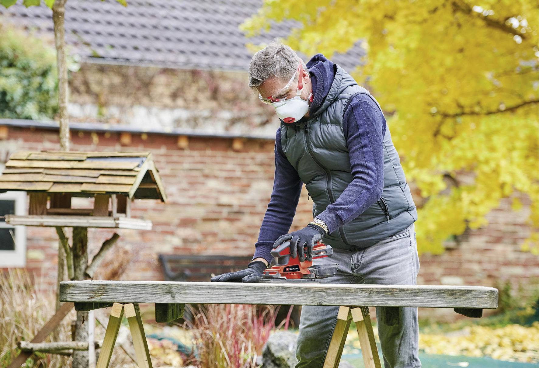 Un homme se tient dehors à une table, portant un masque de protection et des gants, et travaille un morceau de bois avec un outil de meulage électrique.