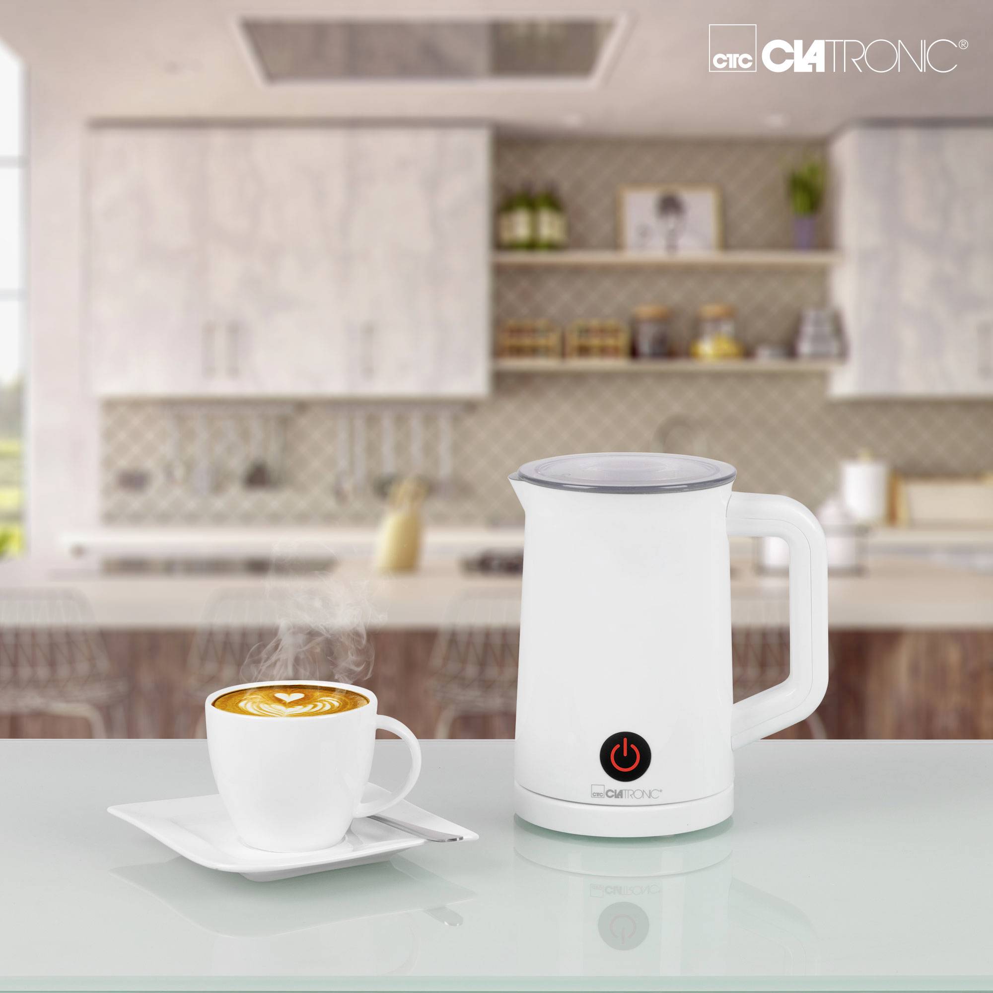 Un mousseur à lait blanc et une tasse de café avec latte art sont posés sur une table dans une cuisine moderne en arrière-plan.