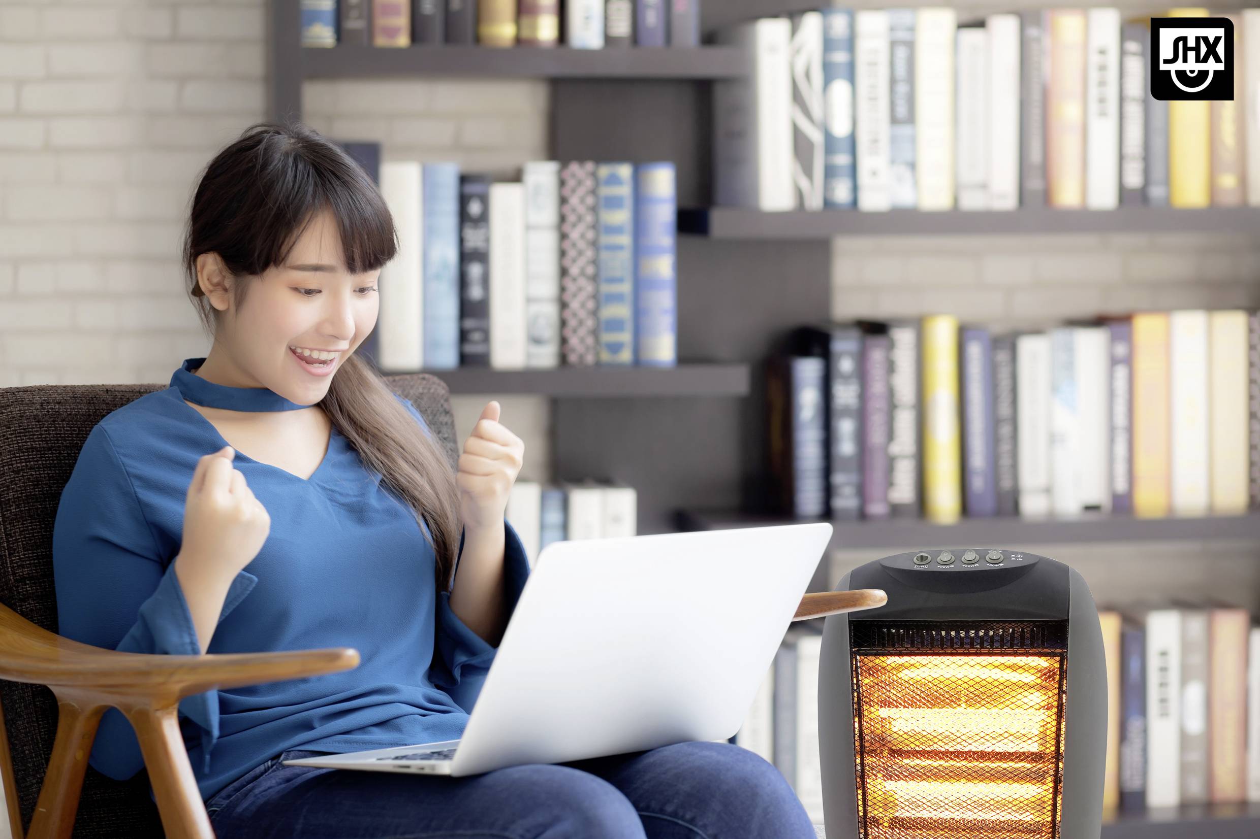 Une femme est assise avec enthousiasme sur un fauteuil, un ordinateur portable sur les genoux. À l'arrière-plan, une bibliothèque est visible et un appareil de chauffage se trouve à droite.