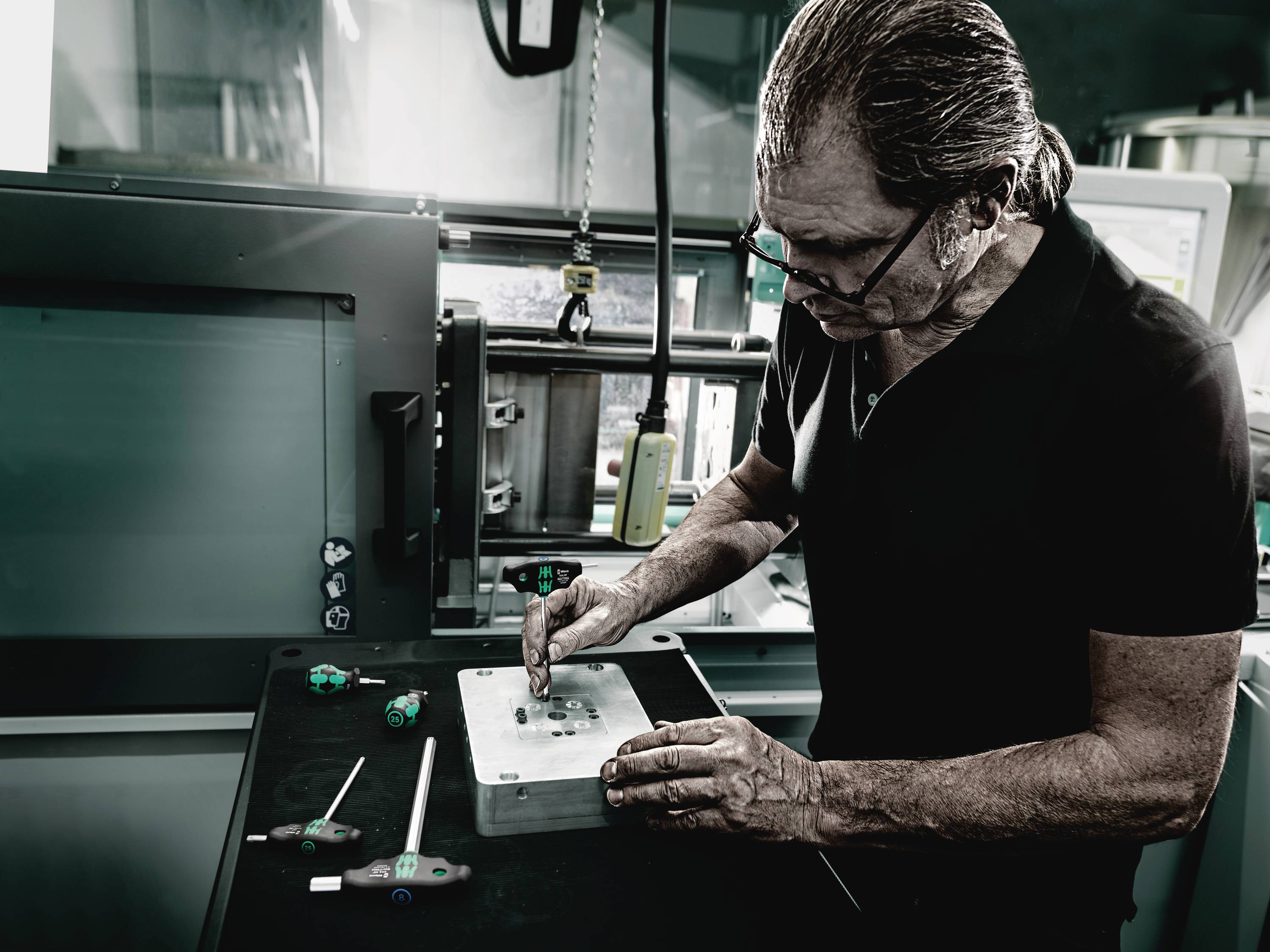 Un homme âgé travaille sur une pièce métallique dans un atelier, entouré d'outils. Il porte un t-shirt noir et des lunettes.