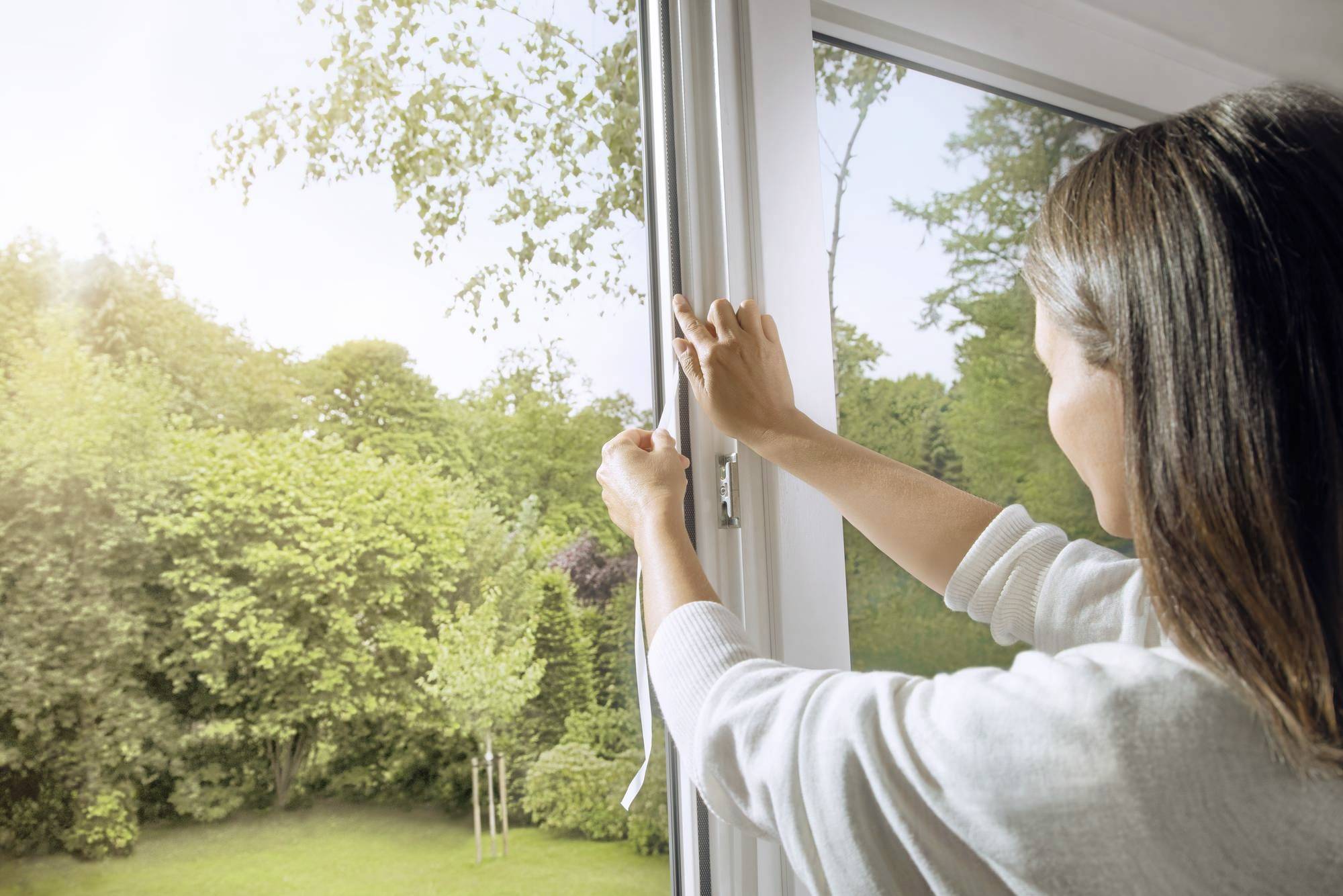 Une femme ouvre une fenêtre pour laisser entrer de l'air frais. À l'extérieur, on aperçoit un jardin verdoyant par une journée ensoleillée.