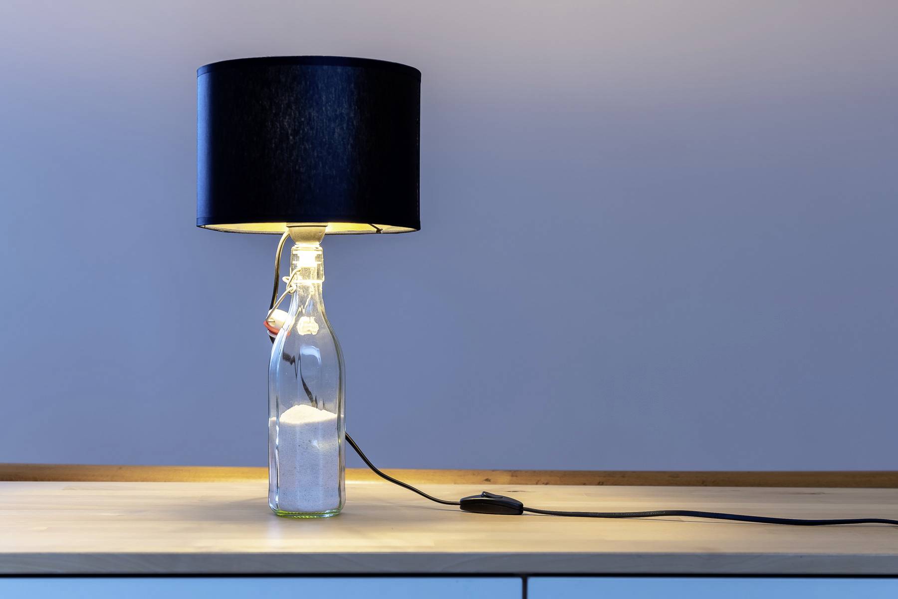 Une lampe de table avec un abat-jour noir, dont le socle est constitué d'une bouteille en verre, est posée sur une table en bois clair.