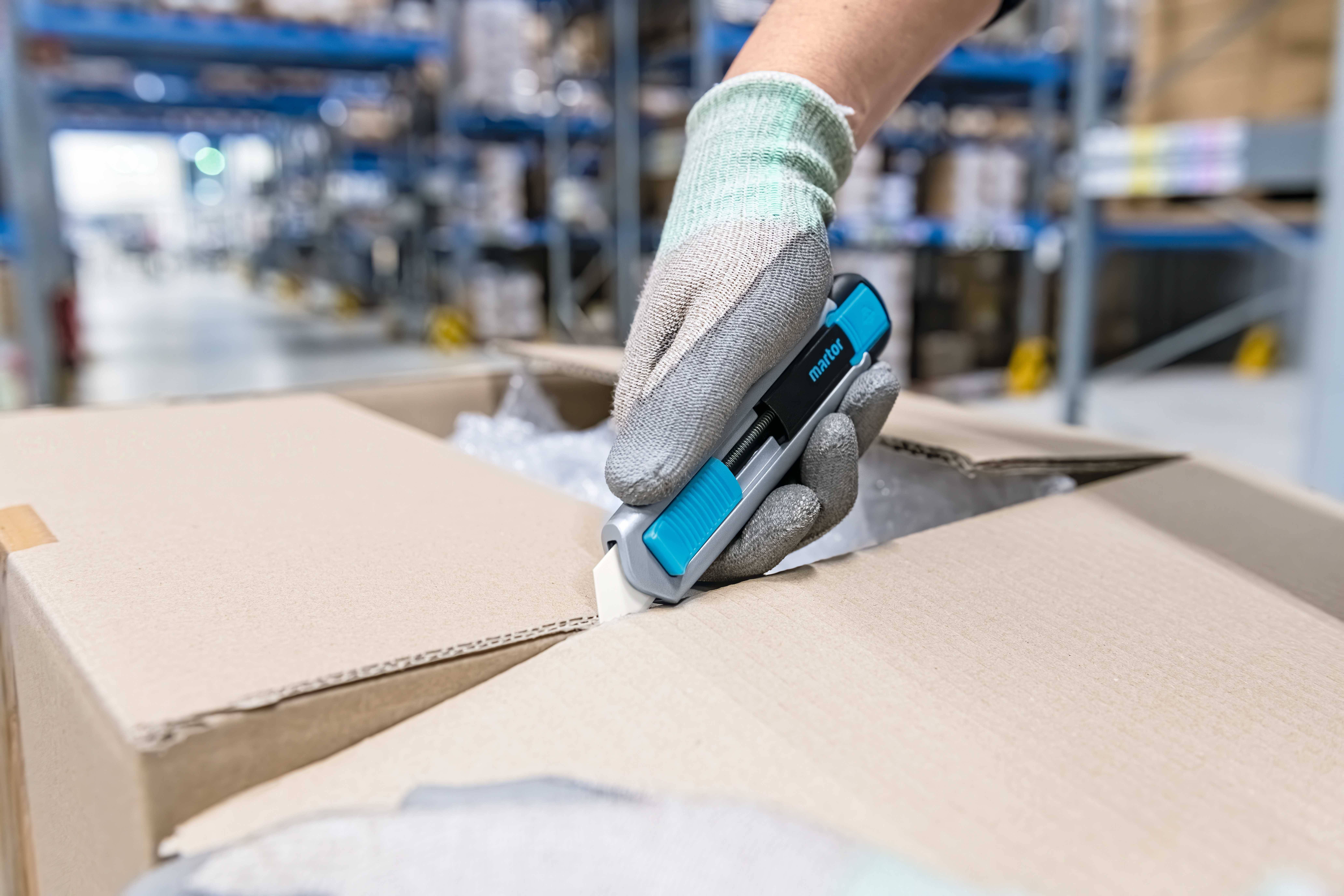 Une personne ouvre un carton avec un couteau de sécurité dans un entrepôt. Des étagères sont visibles en arrière-plan.