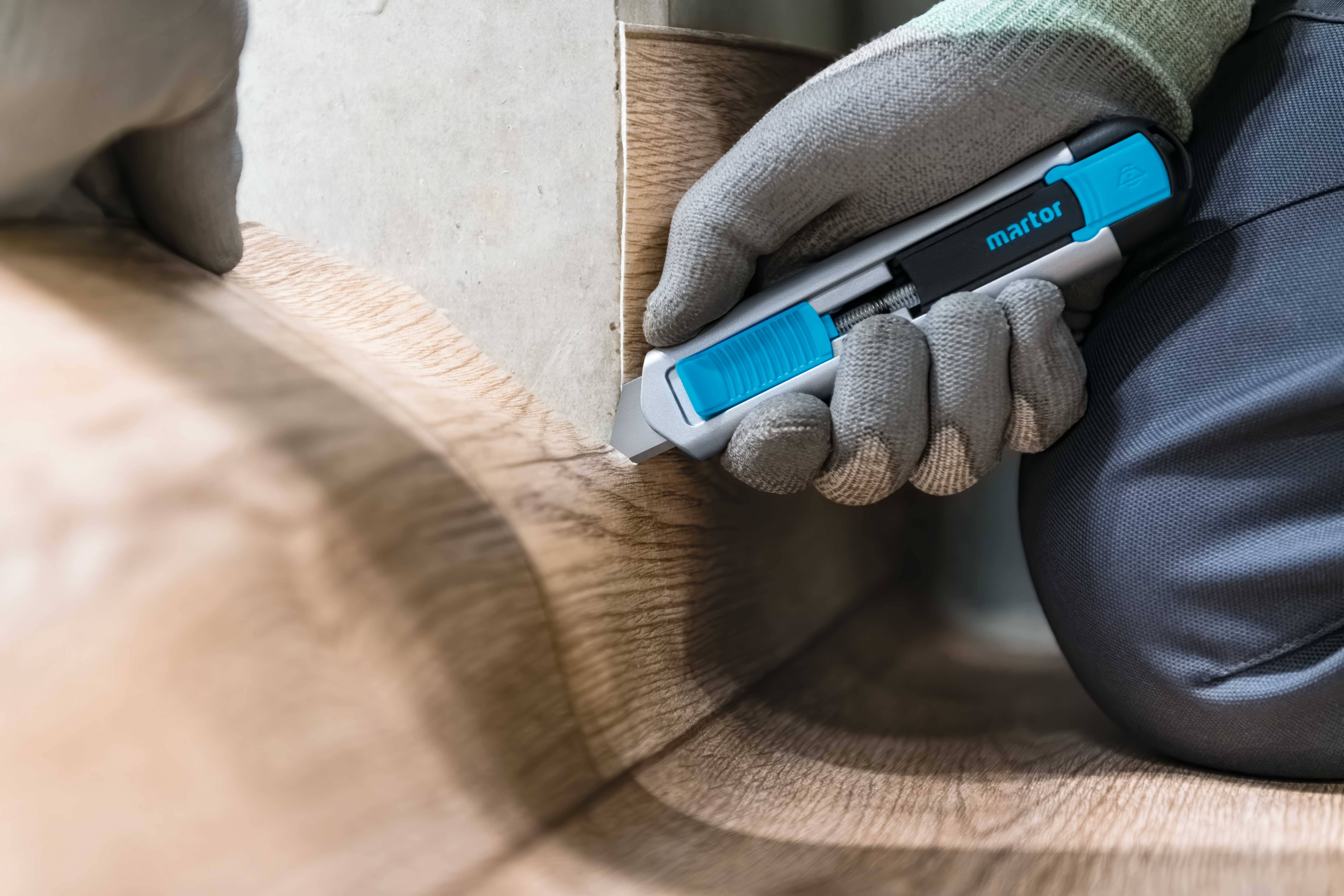 Une personne portant un gant gris utilise un outil de coupe bleu pour découper avec précision un revêtement de sol de type linoléum.