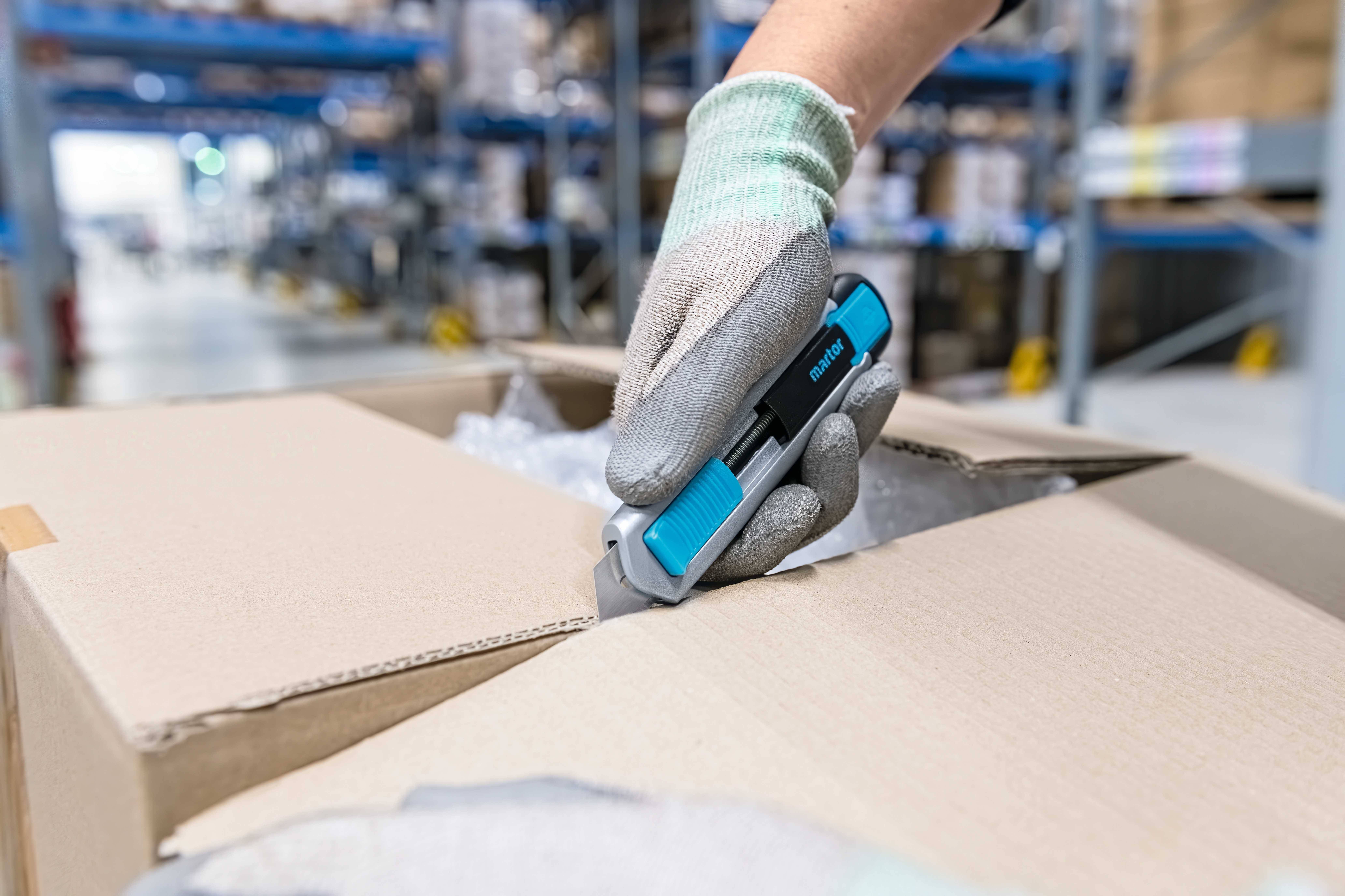Une main gantée ouvre un carton en carton avec un couteau de sécurité bleu dans un entrepôt. Des étagères sont visibles en arrière-plan.