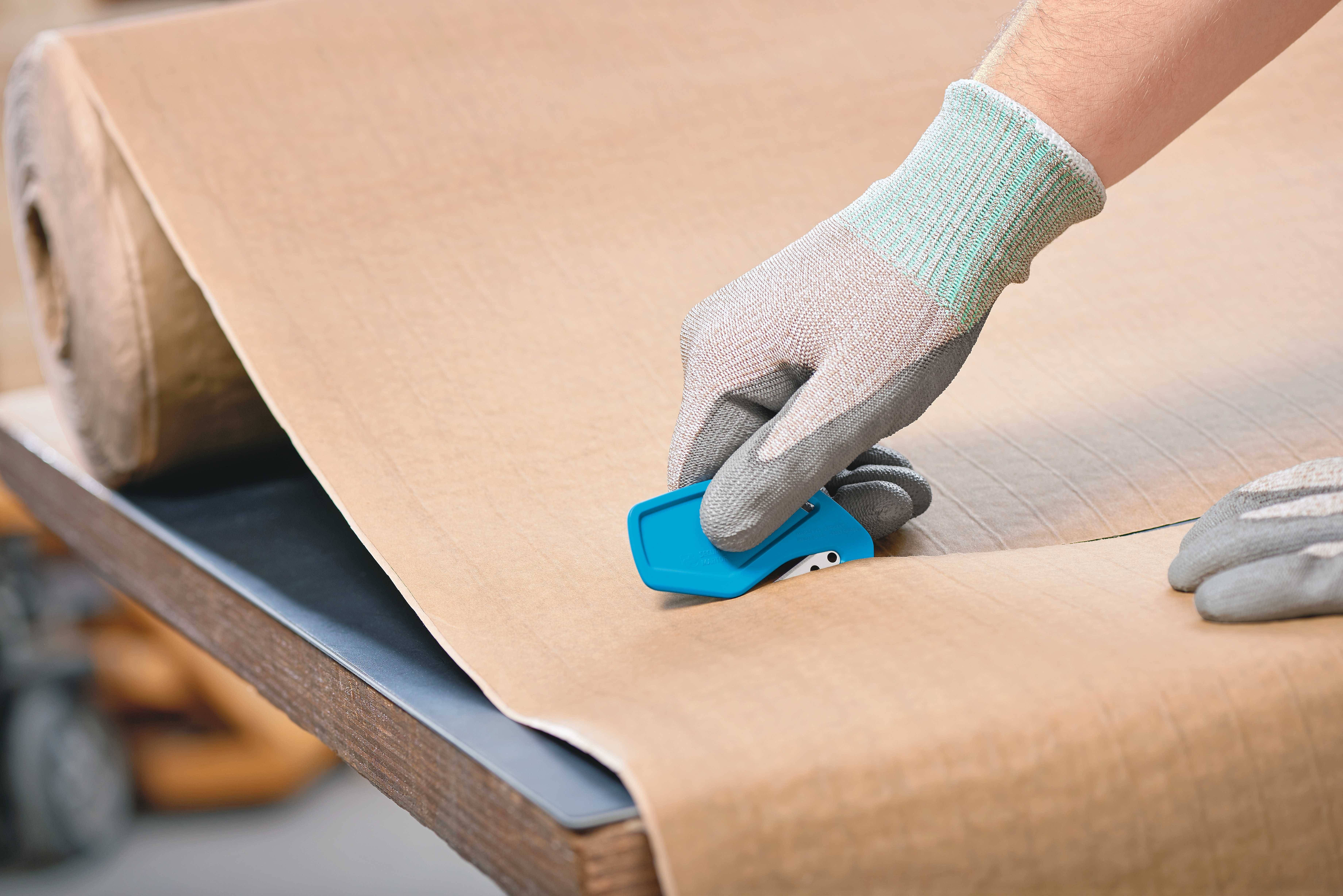Une personne coupe un grand morceau de matériau beige avec un cutter bleu sur une table.