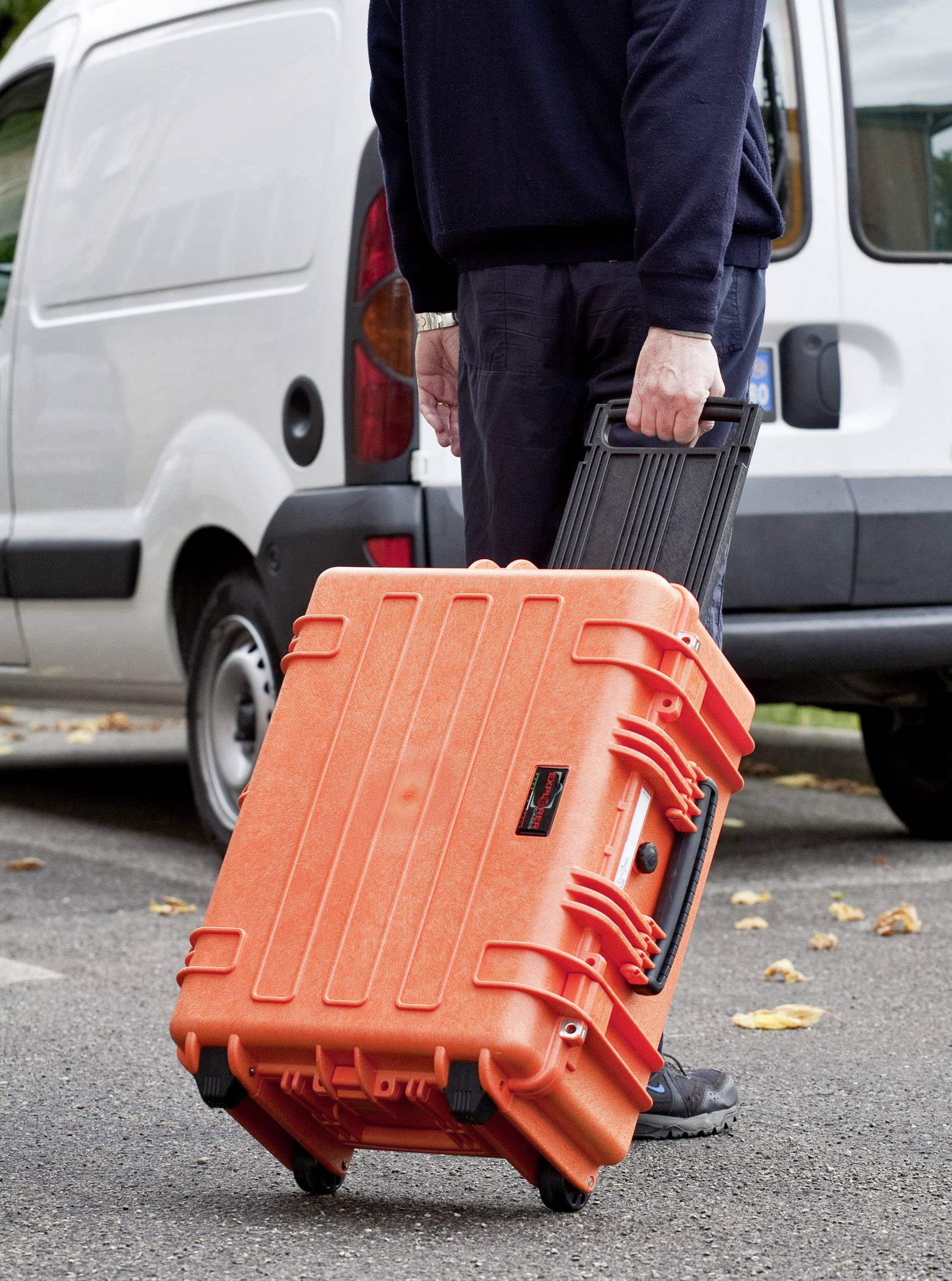 Une personne tire une grande valise orange à roulettes sur un parking à côté d'un véhicule blanc.