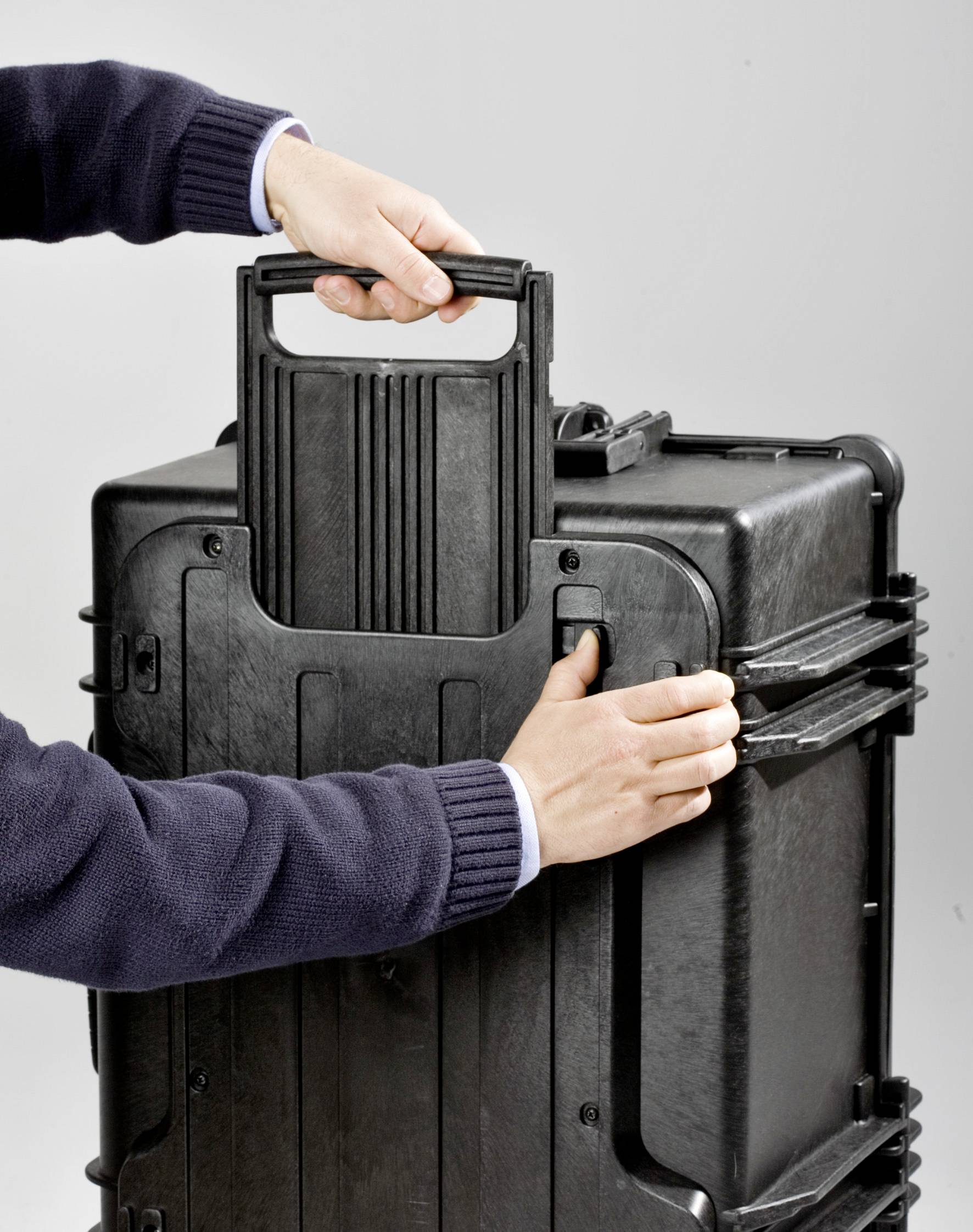 Une personne tire la poignée d'une grande valise noire en plastique. La valise a des coins renforcés et est adaptée au transport.