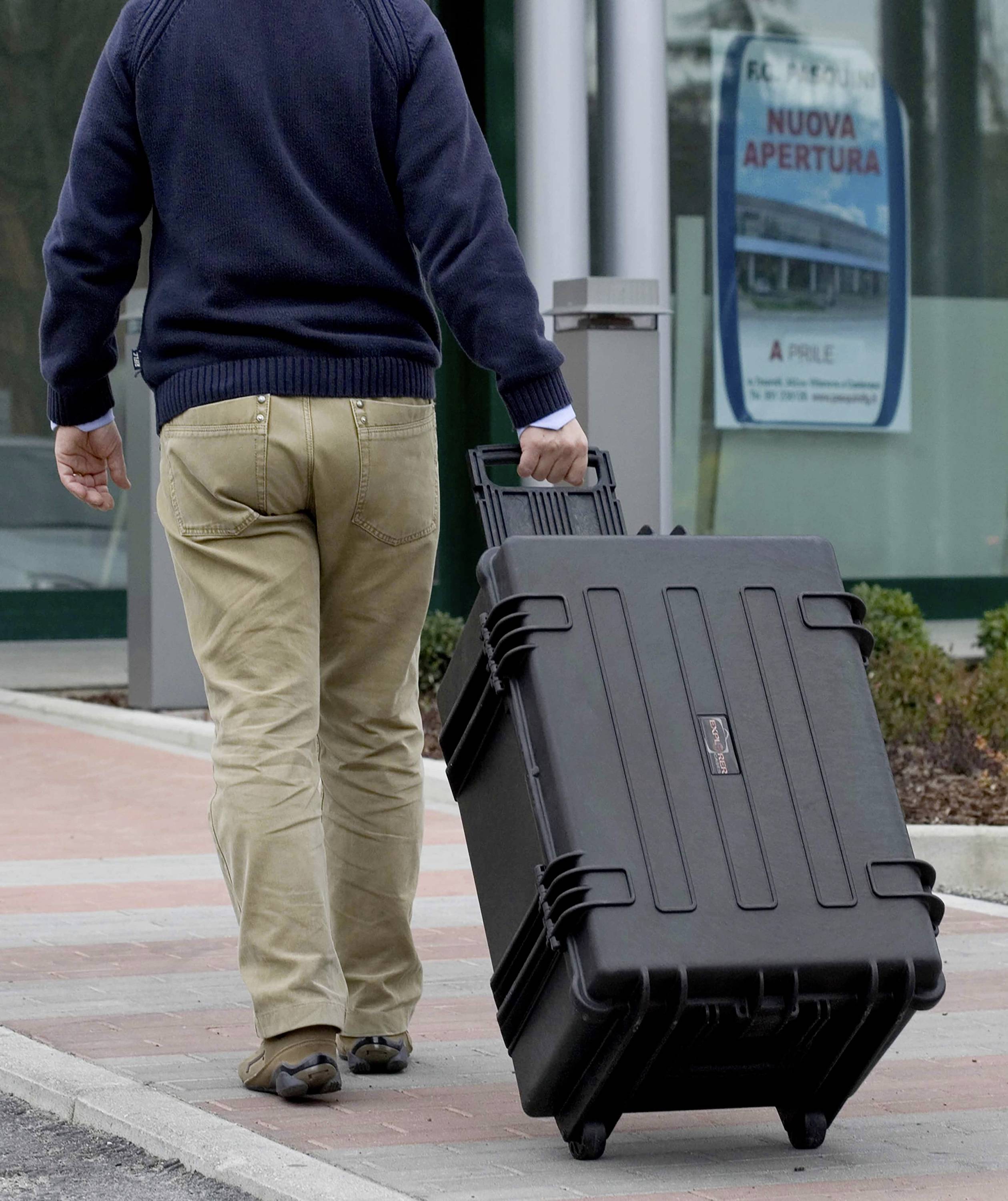 Une personne tire une grande valise noire sur le trottoir. En arrière-plan, un panneau indique « Nuova Apertura ».