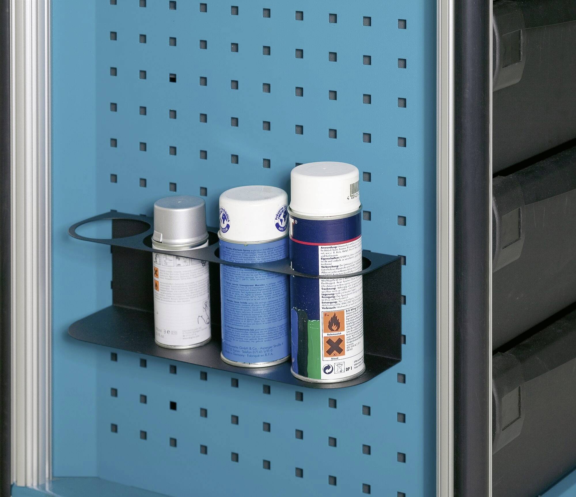 Trois bombes aérosol montées sur une étagère noire devant un mur perforé bleu. À droite, des tiroirs noirs sont visibles.