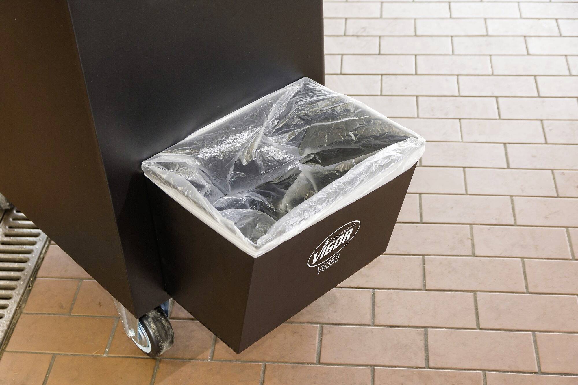 Poubelle noire avec un sac en plastique transparent, montée sur roulettes, posée sur un sol carrelé.