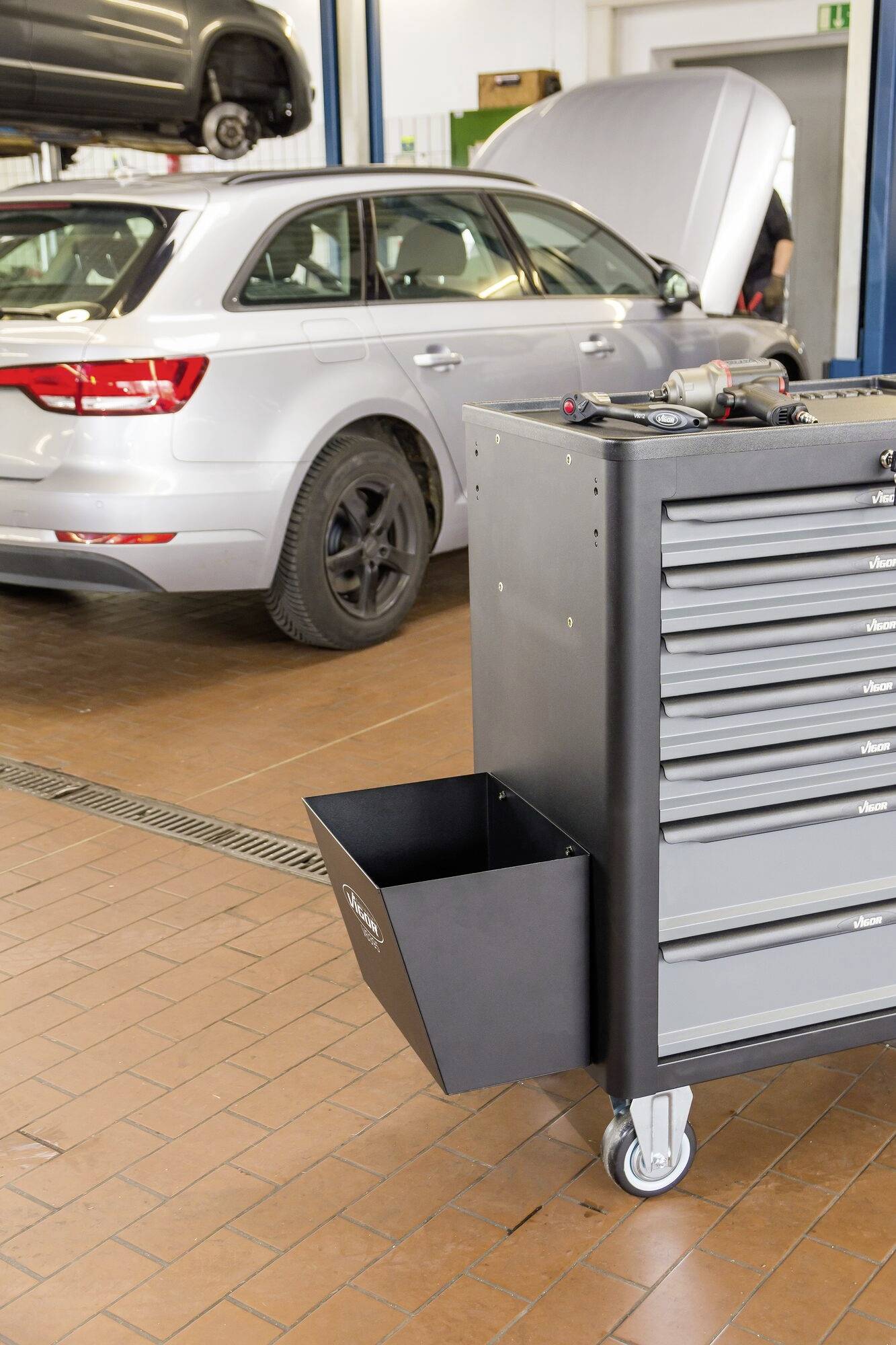 Une voiture grise avec le coffre ouvert est garée dans un atelier. Au premier plan, on peut voir un chariot à outils avec des tiroirs.