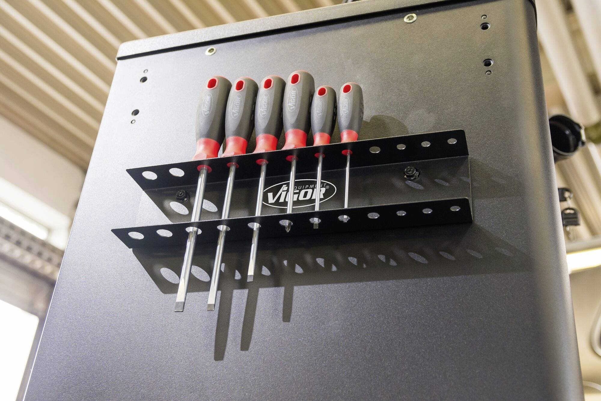 Un ensemble de tournevis avec des poignées rouges est fixé dans un support sur un mur. Au fond, un plafond à lamelles est visible.