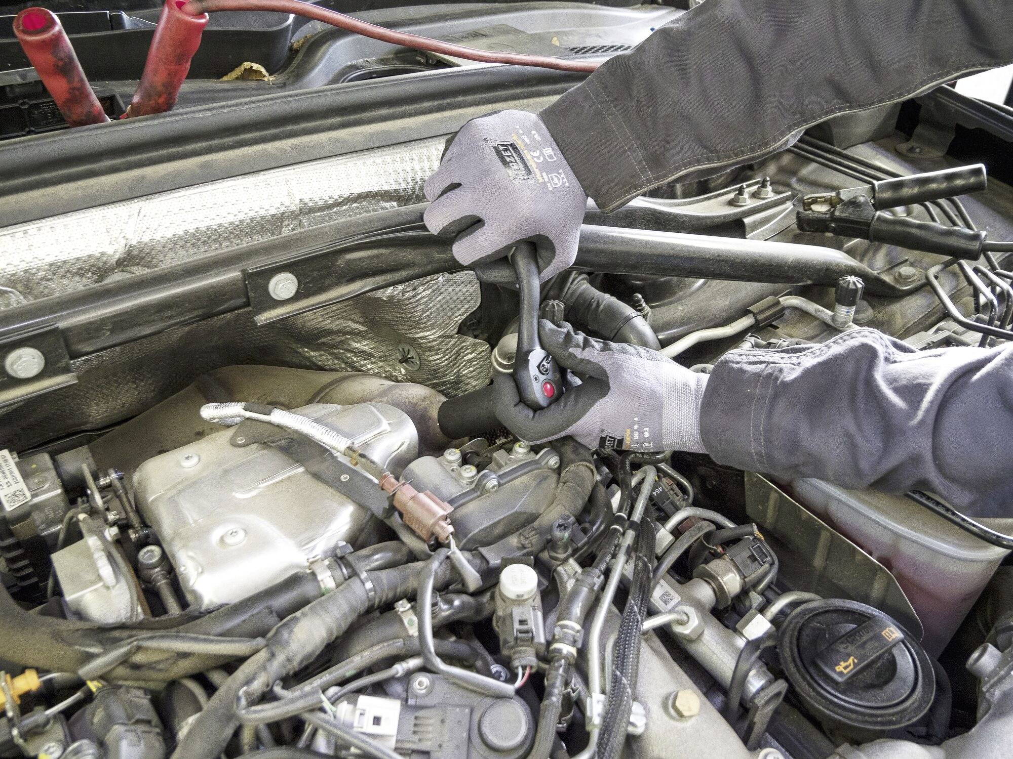 Une personne répare un moteur dans une voiture. Elle tient un outil et porte des gants en caoutchouc. Le compartiment moteur est visible en détail.