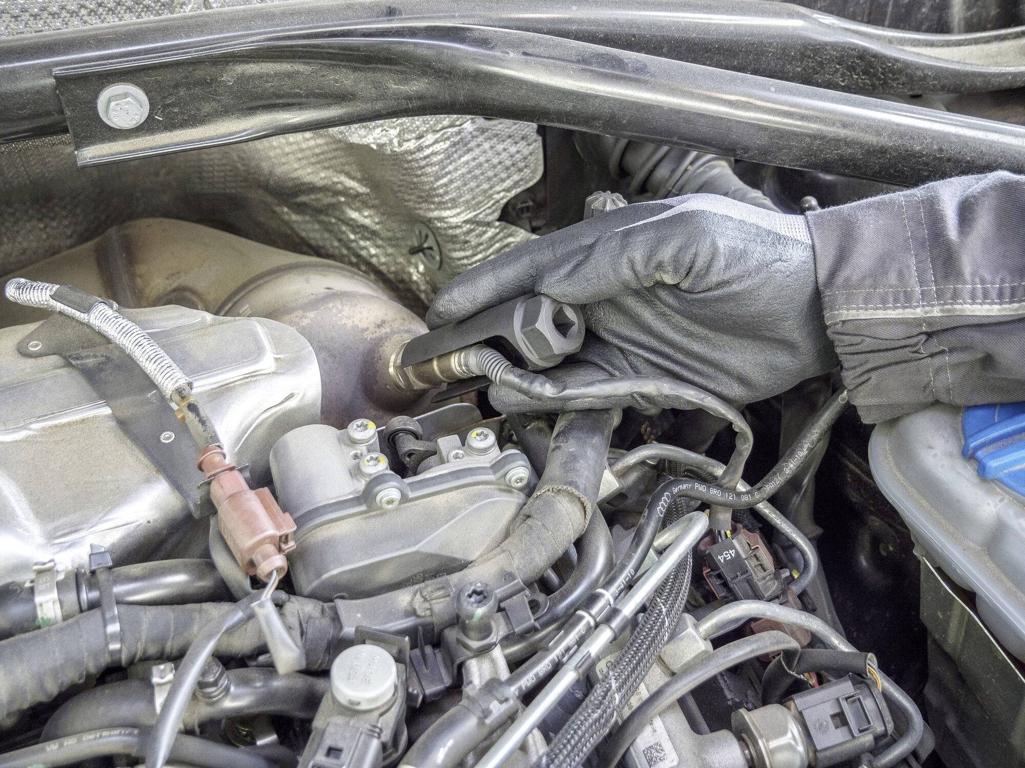 Une main gantée retire une bougie d'allumage du compartiment moteur d'une voiture. Les environs du moteur et les pièces adjacentes sont visibles.