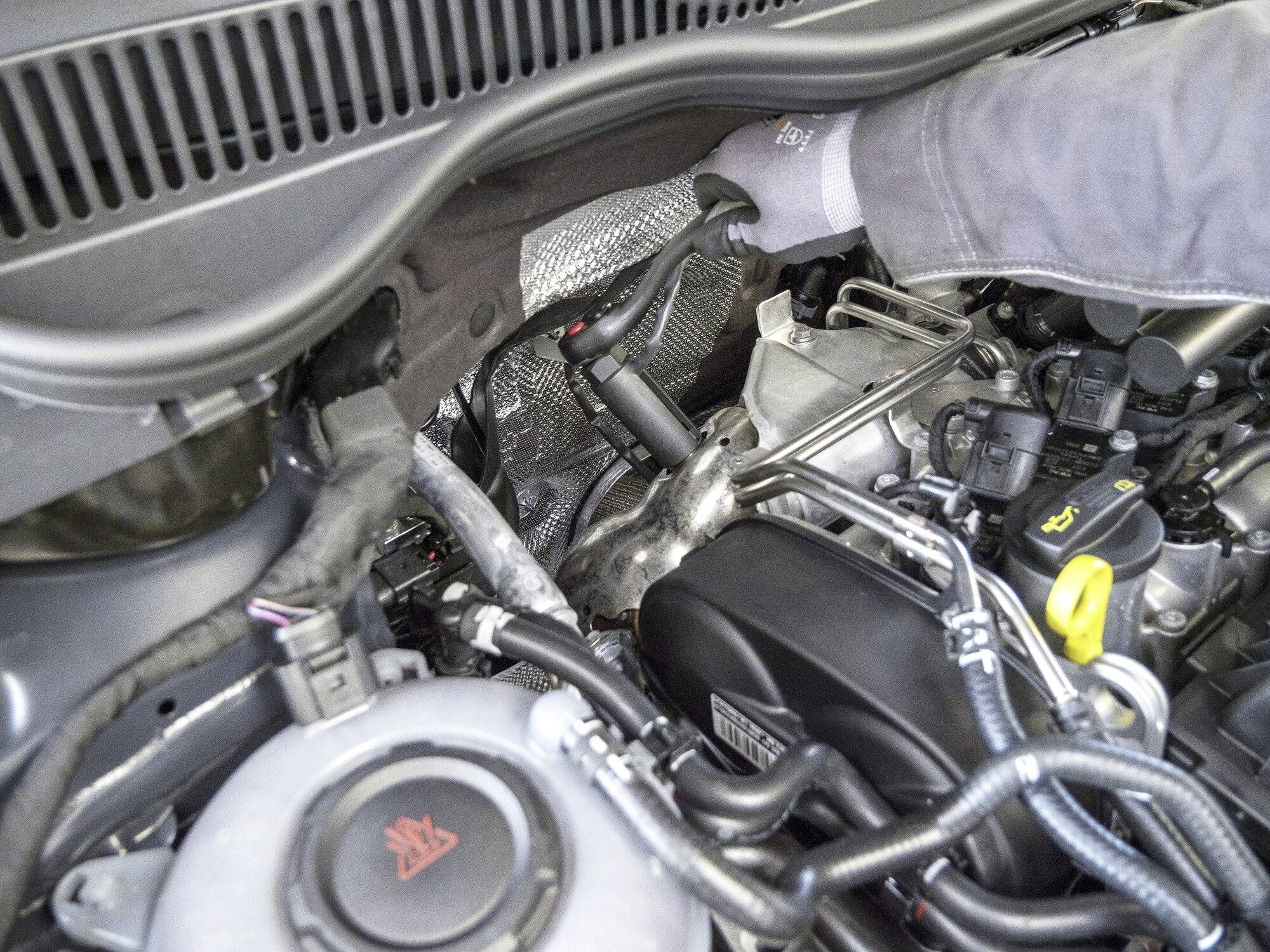 Une personne vérifie les connexions du moteur dans une voiture. Divers câbles et composants moteur sont visibles sous le capot.