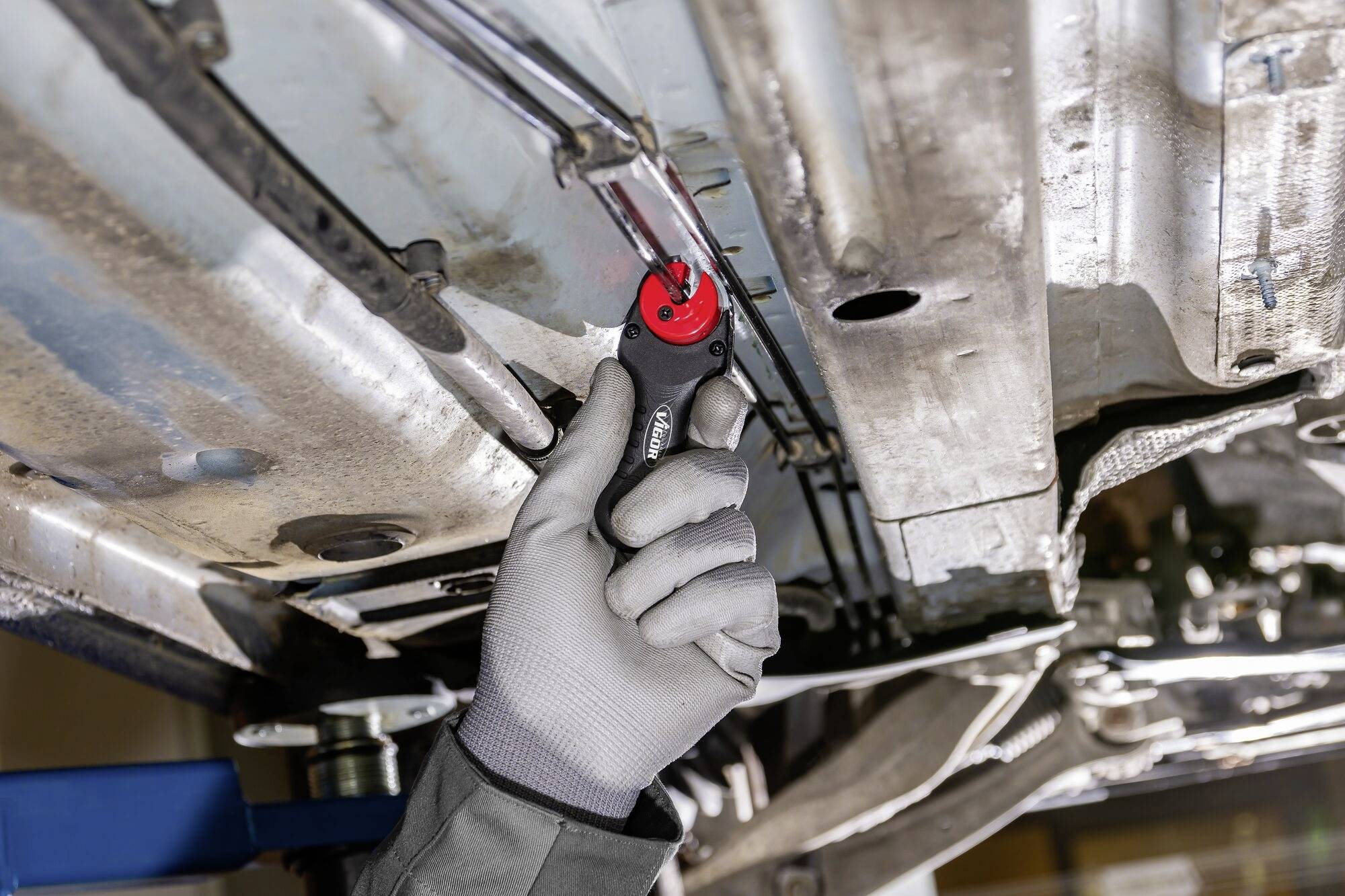 Un mécanicien portant des gants gris utilise un outil rouge pour travailler sous un véhicule.