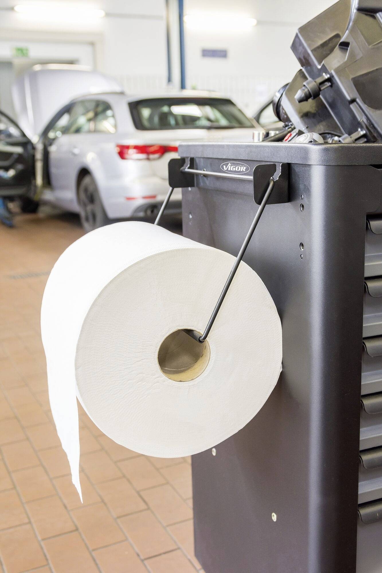 Un grand rouleau de papier est suspendu à un chariot à outils dans un atelier automobile. À l'arrière-plan, une voiture est stationnée avec son capot ouvert.