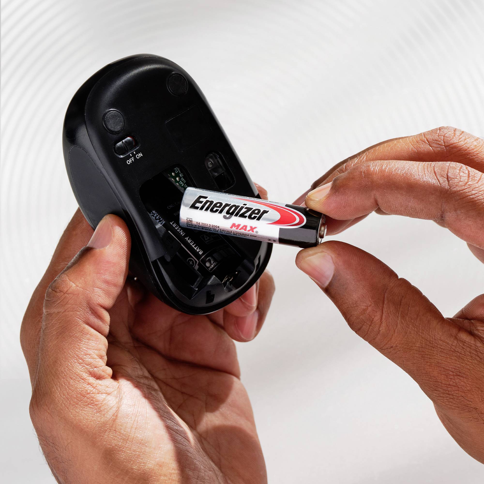 Deux mains tiennent une souris d'ordinateur pendant qu'une pile est insérée. La pile arbore le logo « Energizer Max ».