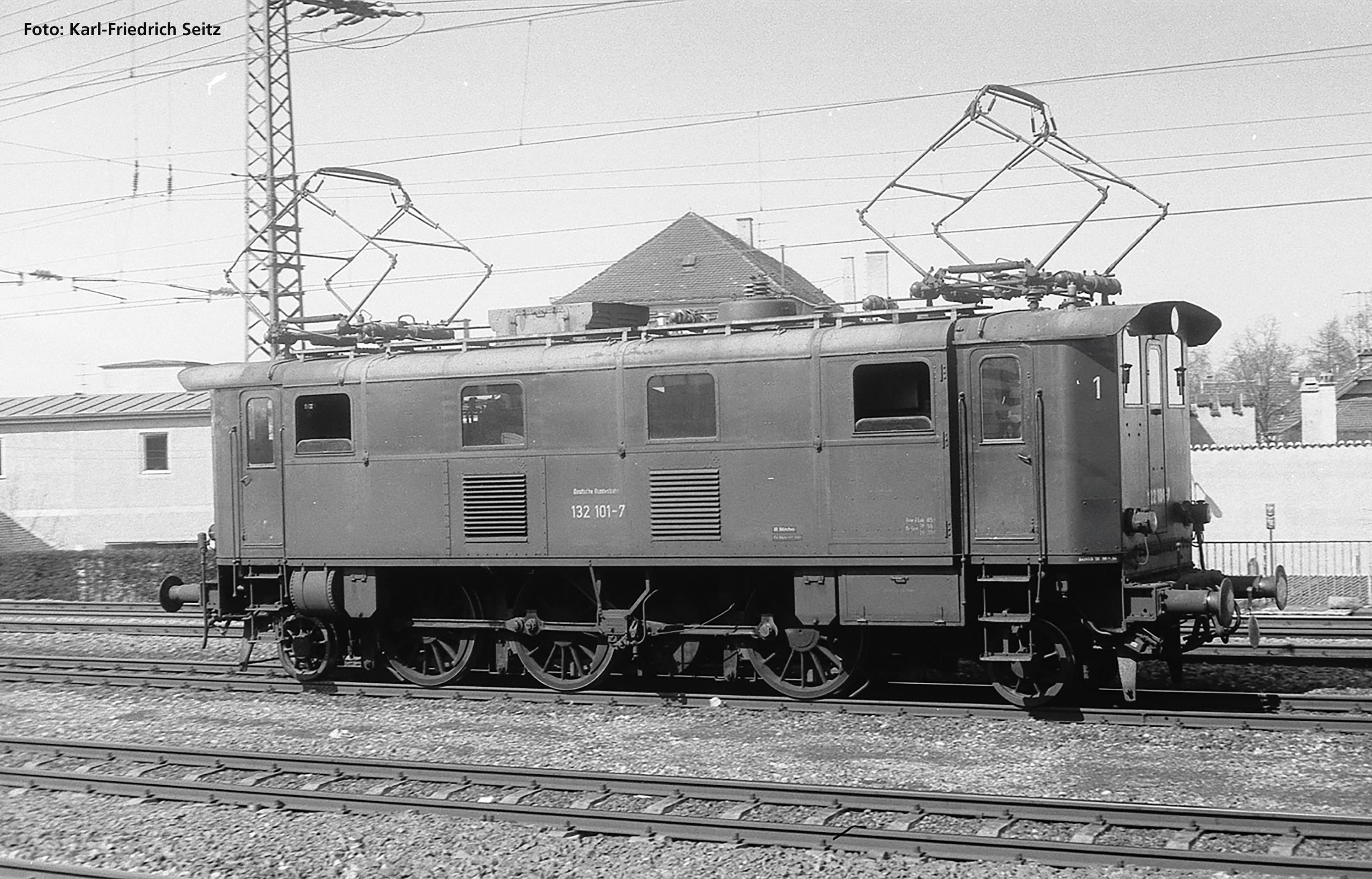 Une photographie en noir et blanc d'une locomotive électrique historique sur des voies ferrées. Des lignes électriques sont visibles en arrière-plan.