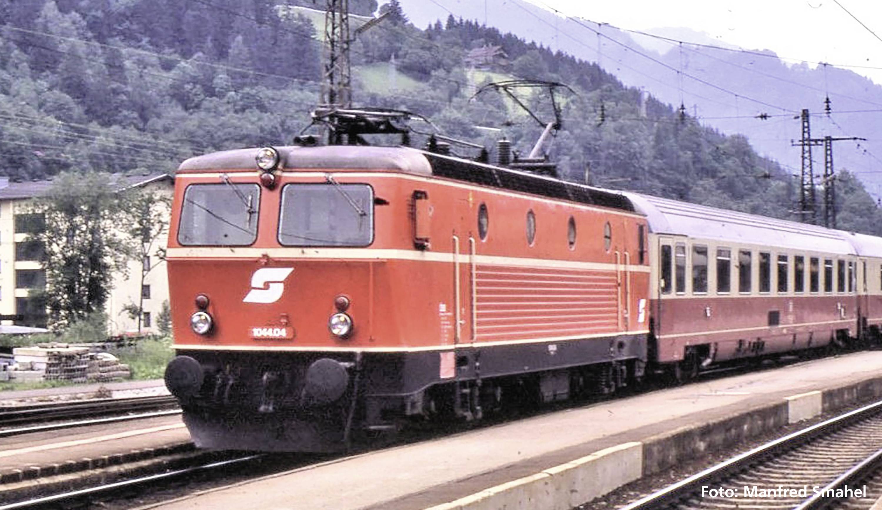 Un train rouge portant l'inscription 'S' traverse une gare rurale, avec des montagnes et des forêts en arrière-plan.