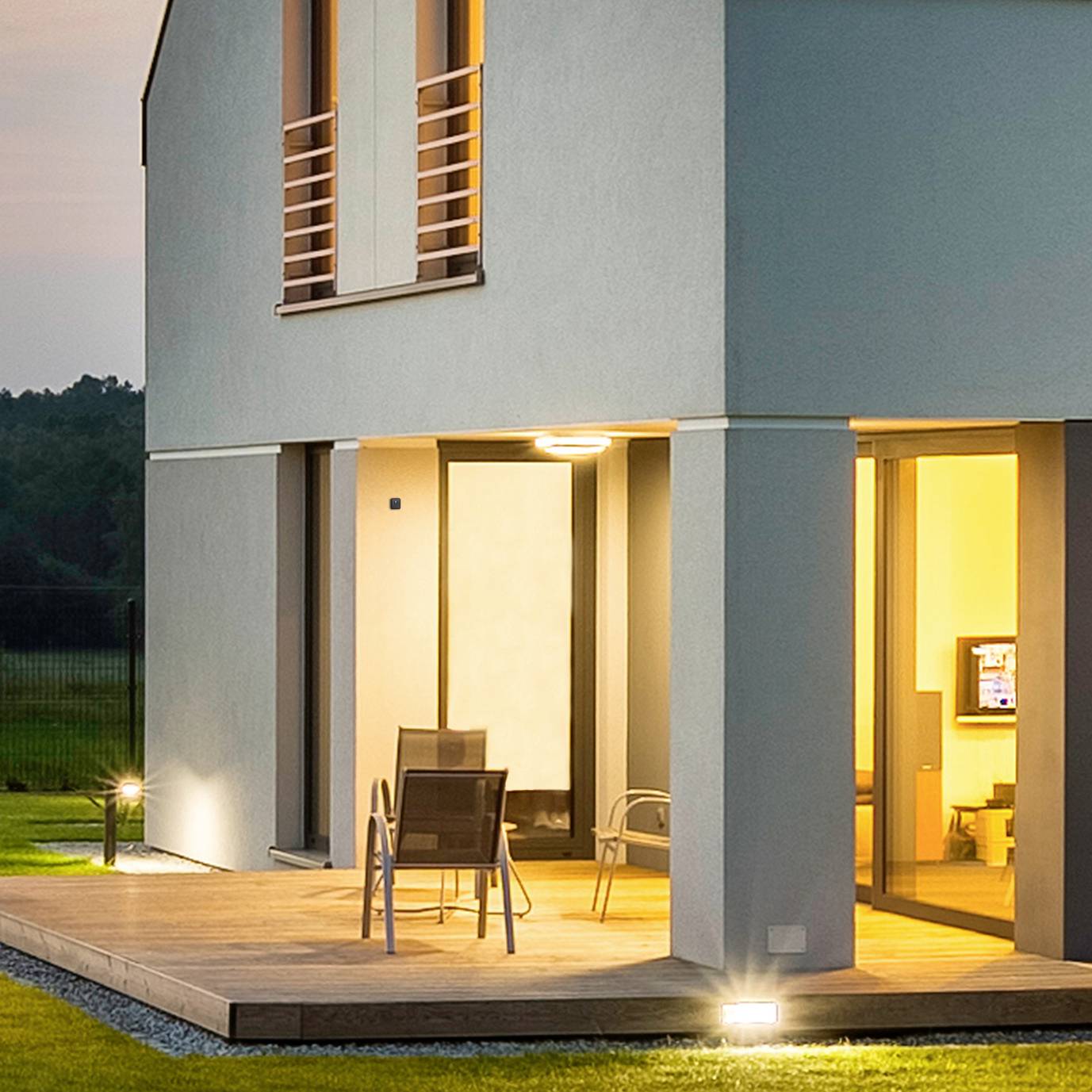 Maison moderne à deux étages au crépuscule. L'éclairage extérieur illumine le hall d'entrée et la terrasse, où des chaises sont disposées.