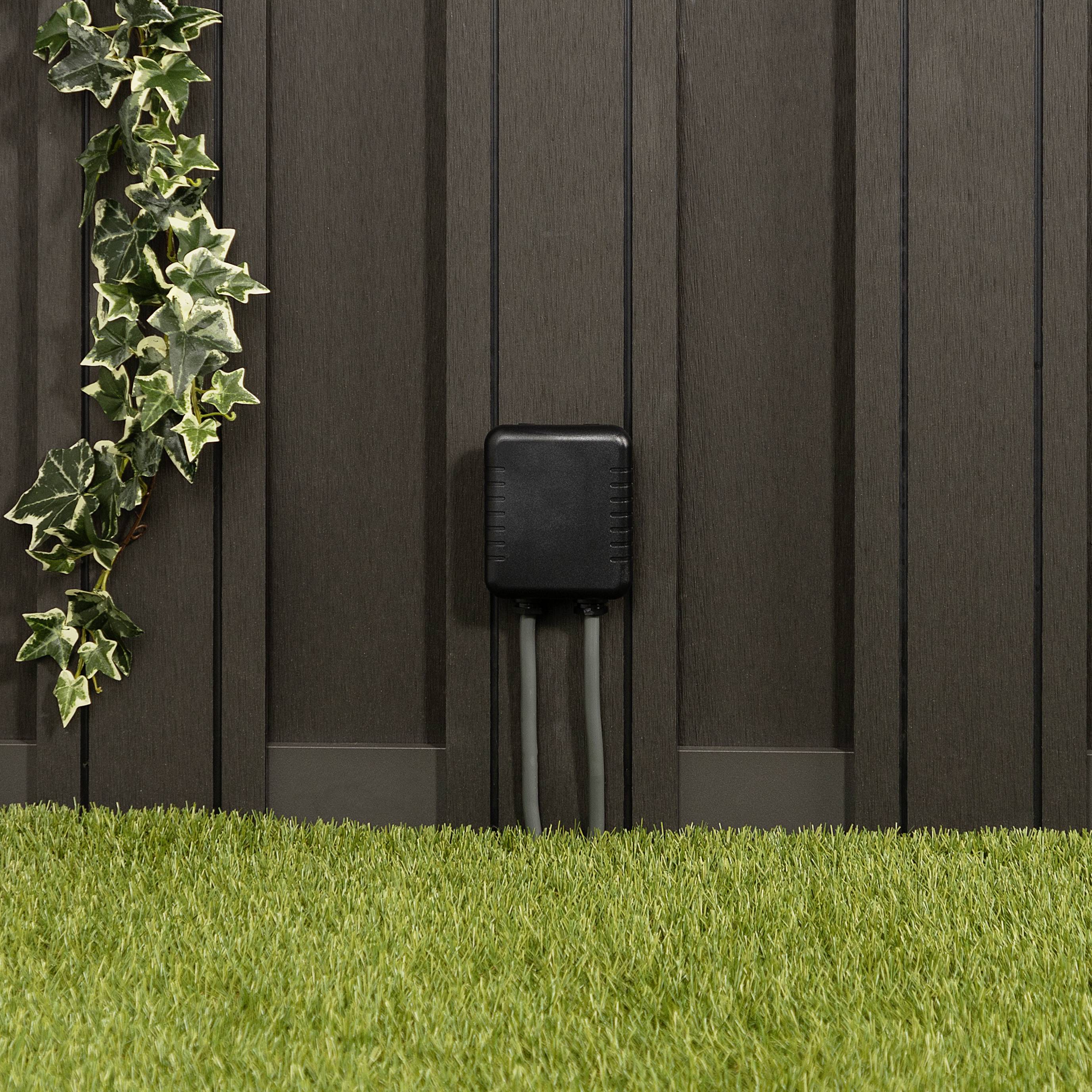 Un appareil électrique avec trois câbles est fixé à un mur moderne. Une plante grimpante est suspendue à côté à gauche. Devant se trouve une surface de pelouse verte.