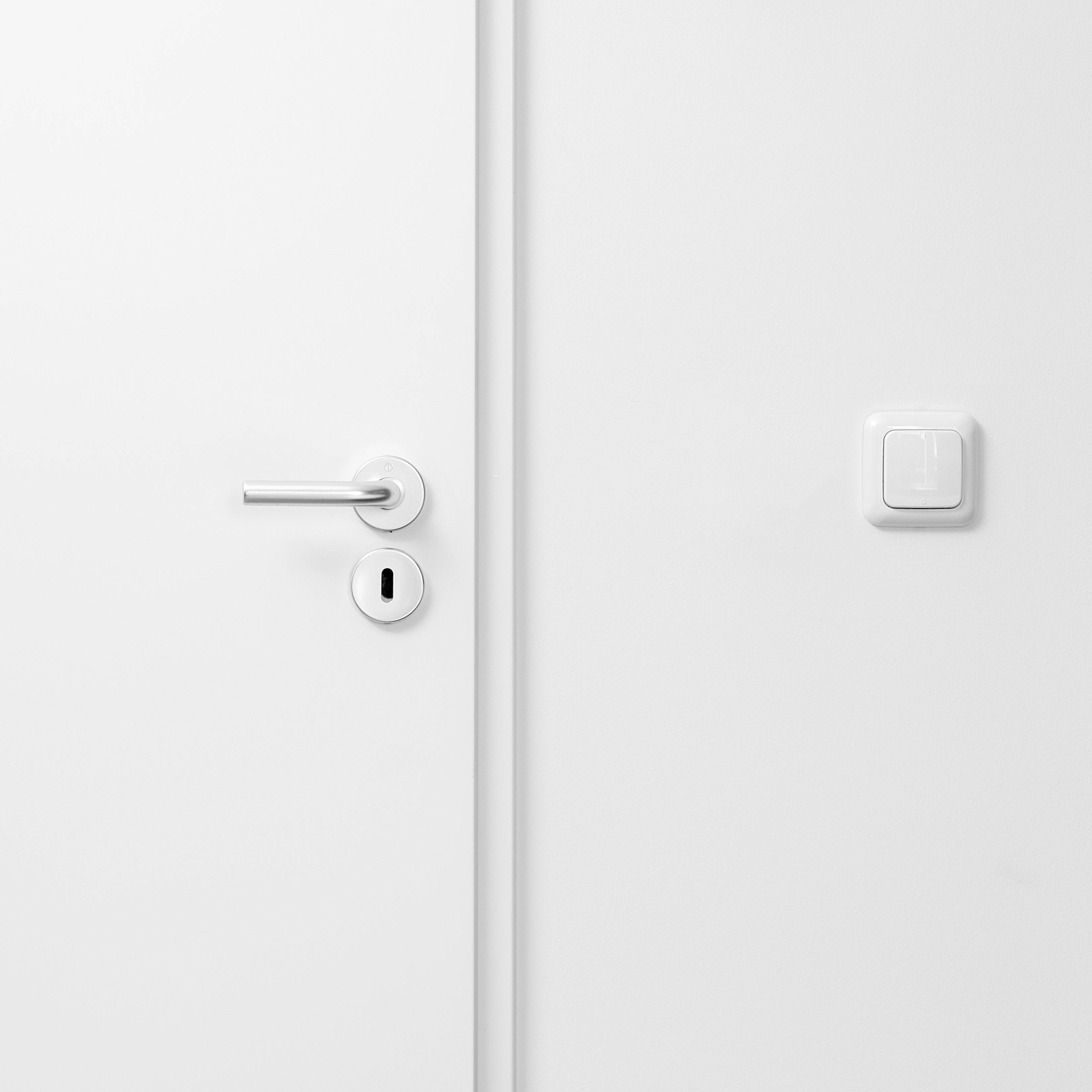 Porte blanche avec poignée argentée et trou de serrure sur le côté gauche ; interrupteur à droite de la porte. Design minimaliste.