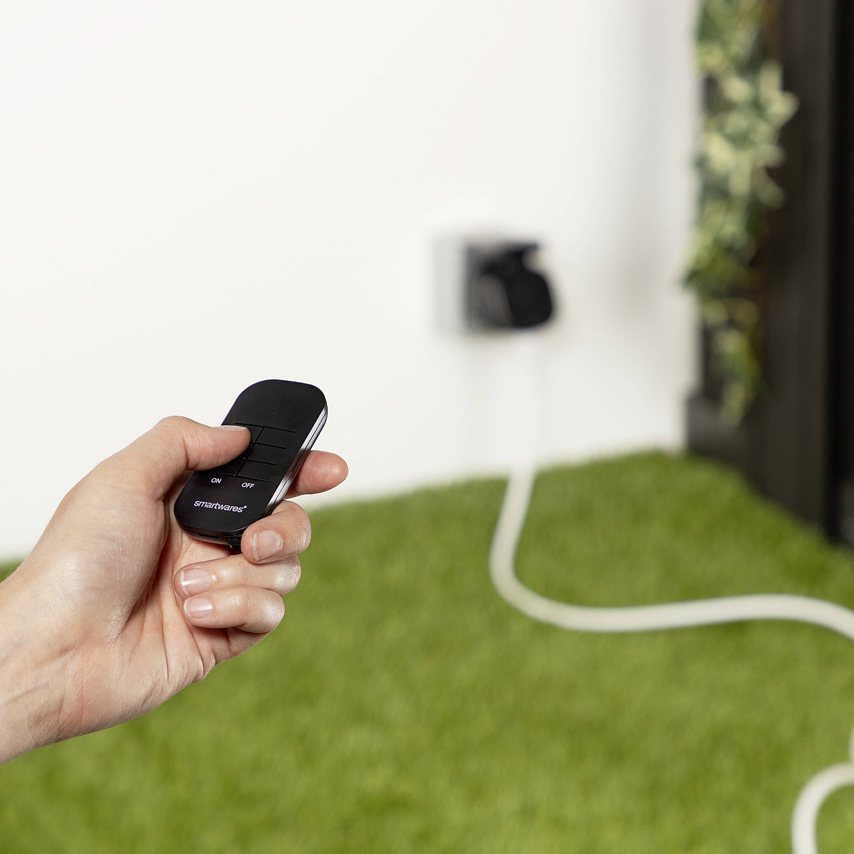 Une main tient une télécommande dans une pièce avec une prise électrique en arrière-plan. Un câble de rallonge est visible.
