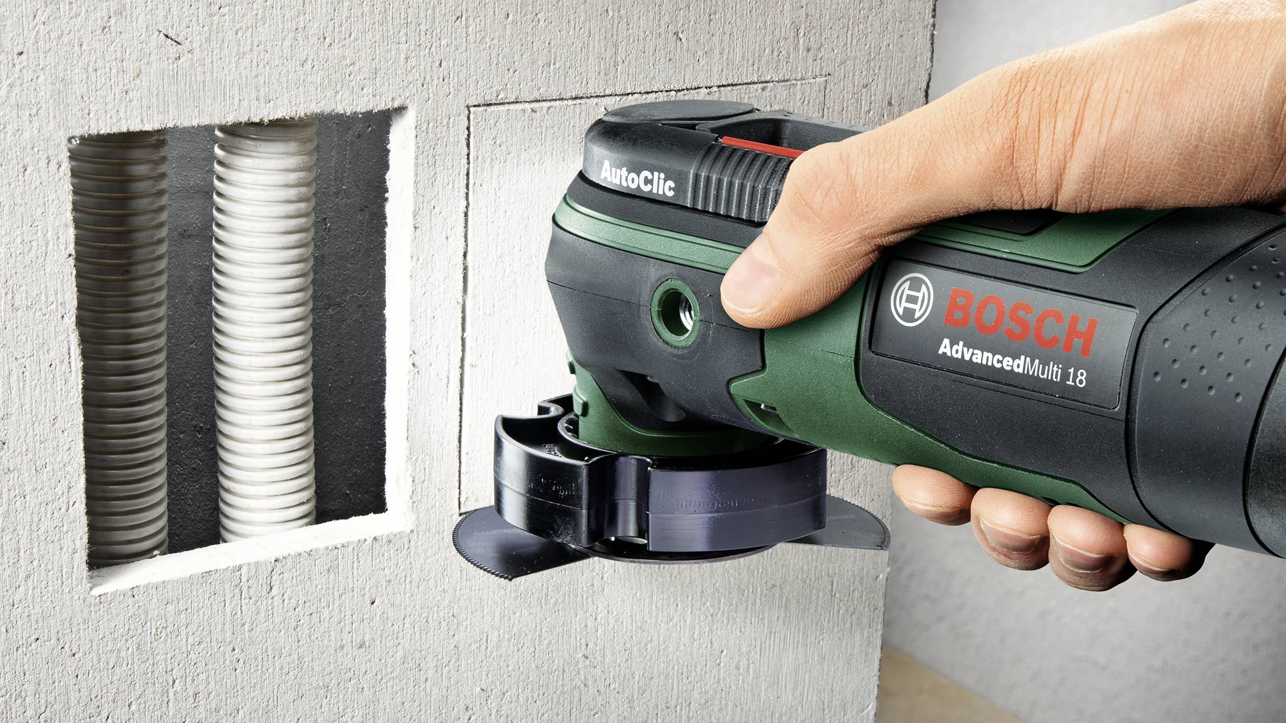Une personne utilise un outil électrique vert pour découper un trou carré dans un mur gris. Des câbles sont visibles à droite.