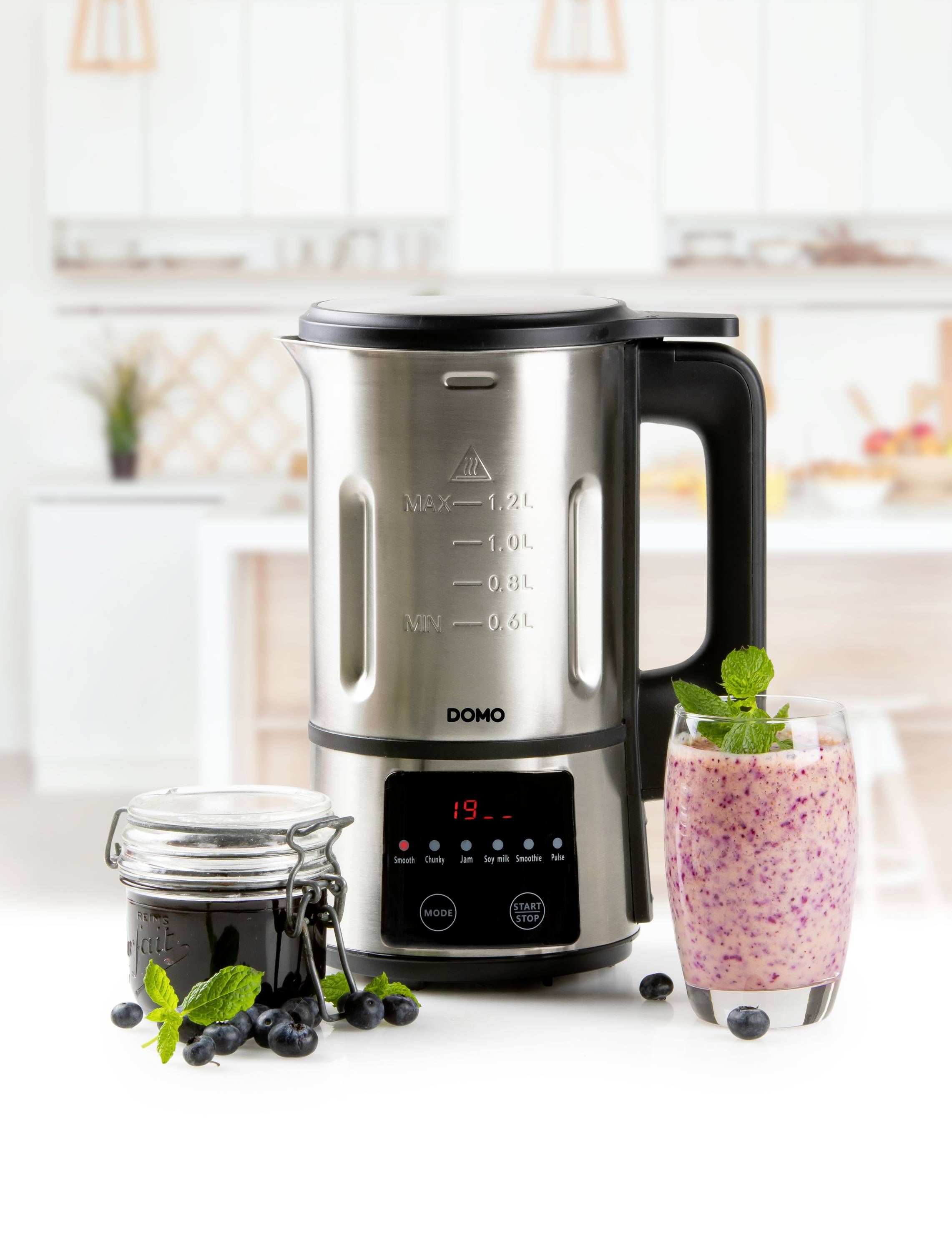 Un mixeur est posé sur un plan de travail de cuisine avec un verre de smoothie et un verre de confiture à côté. Une cuisine est visible en arrière-plan.