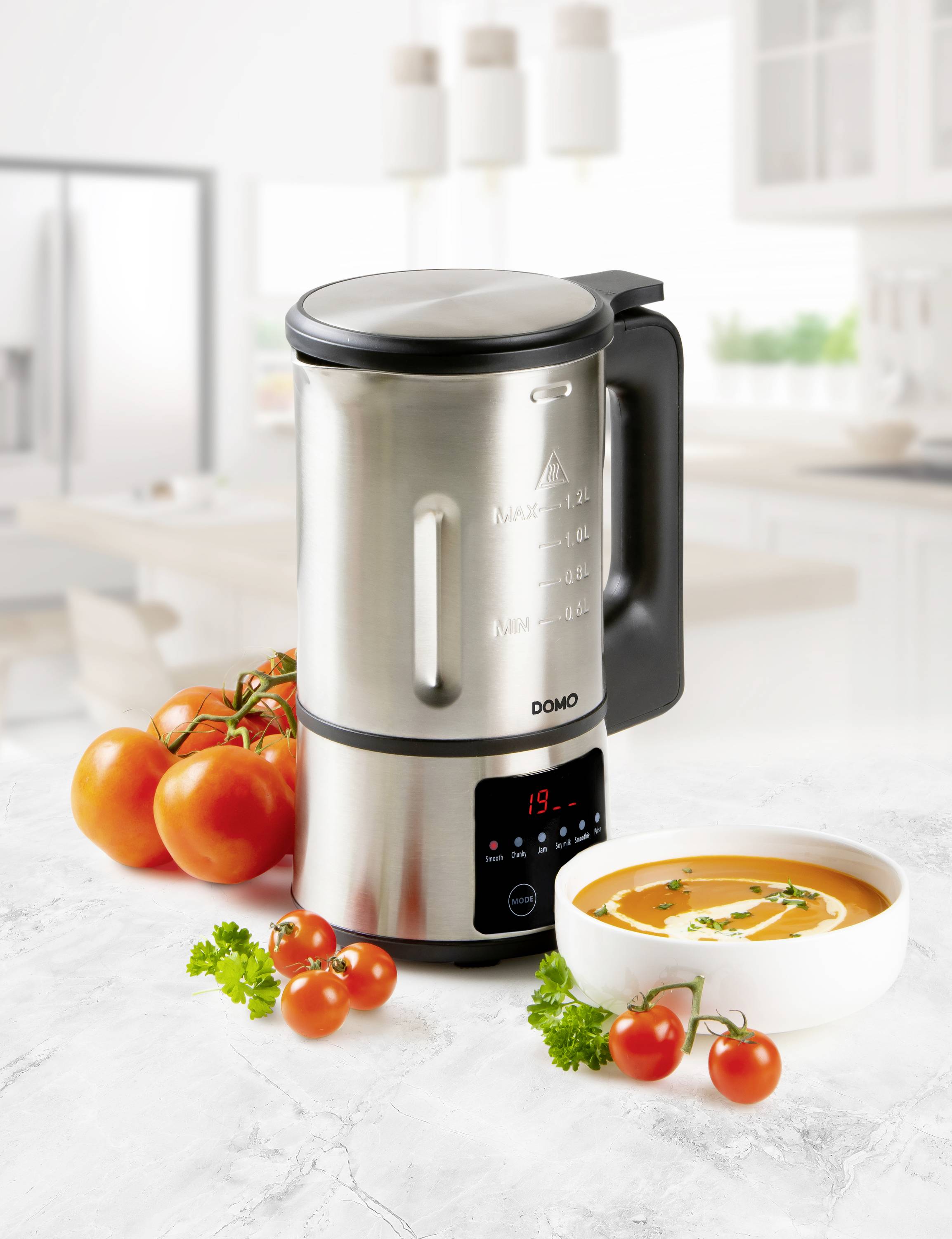 Un préparateur de soupe moderne en acier inoxydable est posé sur un plan de travail de cuisine. À côté se trouvent des tomates, et un bol de soupe est servi.