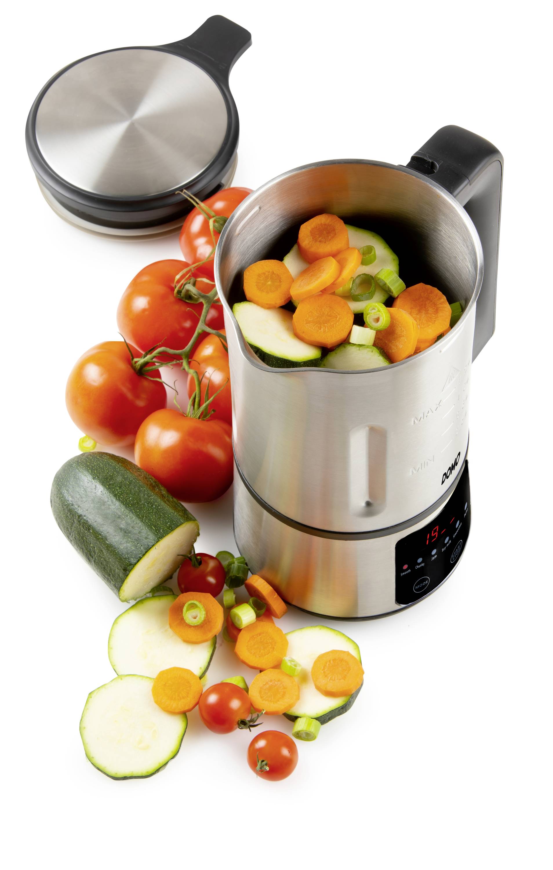 Un mixeur en acier inoxydable rempli de légumes coupés comme des carottes et des courgettes. À côté, des tomates fraîches sont disposées.