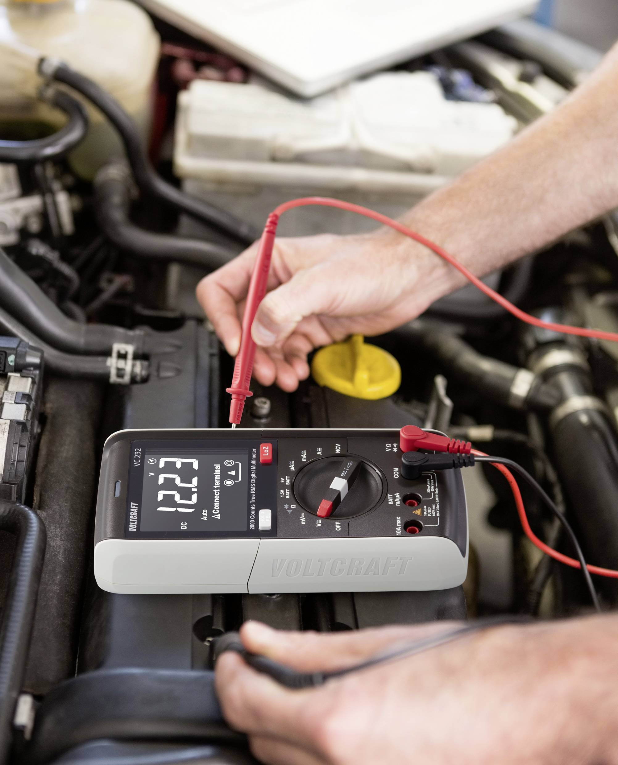 Une personne mesure la tension de la batterie d'une voiture avec un multimètre numérique qui indique 12,23 volts. Les câbles sont connectés.