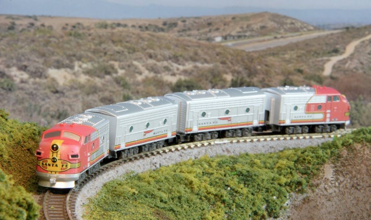 Un train miniature aux couleurs rouge et argentée circule sur un rail sinueux à travers un paysage vallonné et verdoyant.