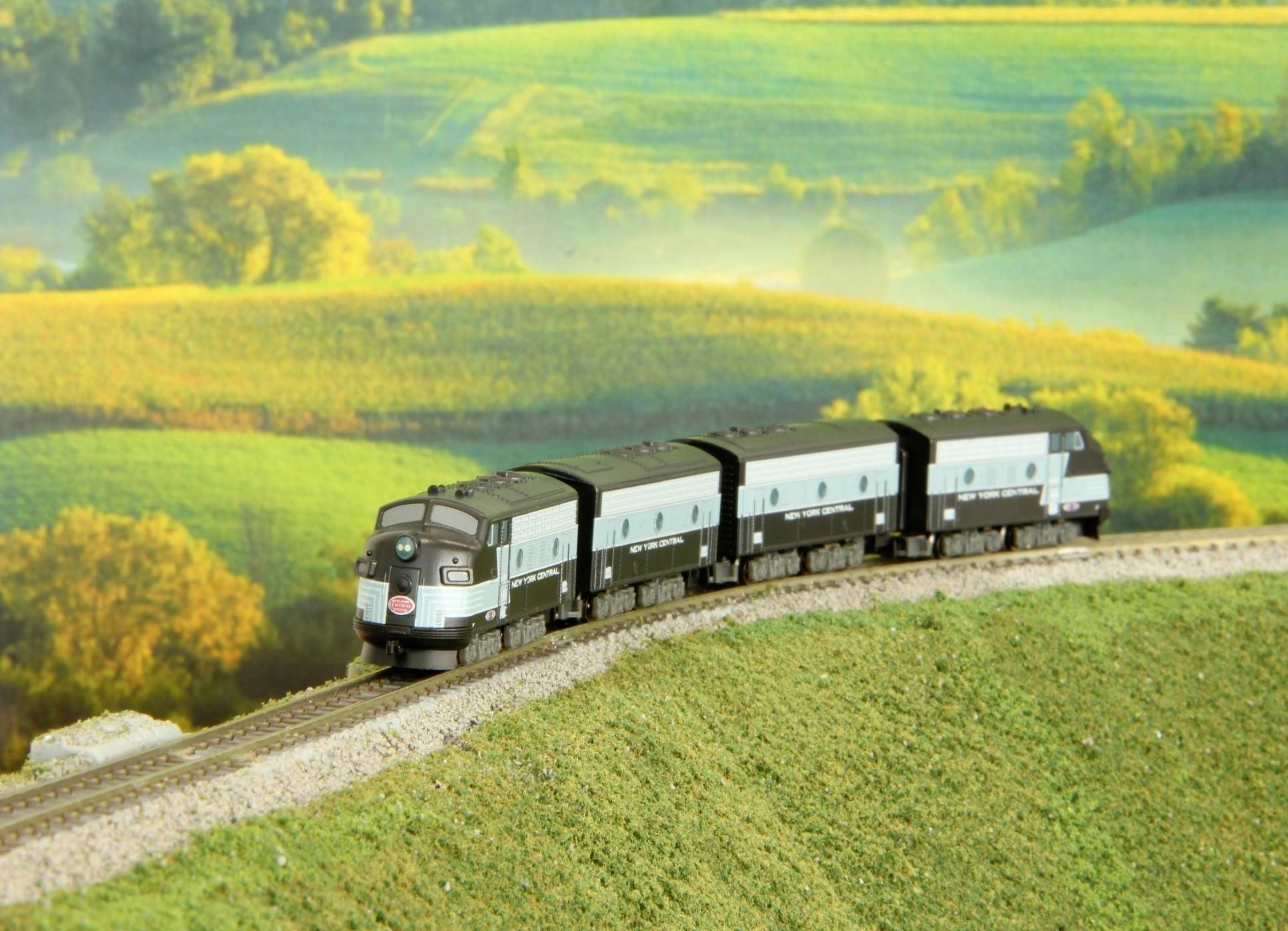 Un train traverse un paysage verdoyant et vallonné. En arrière-plan, on aperçoit des champs en pente douce et des arbres.