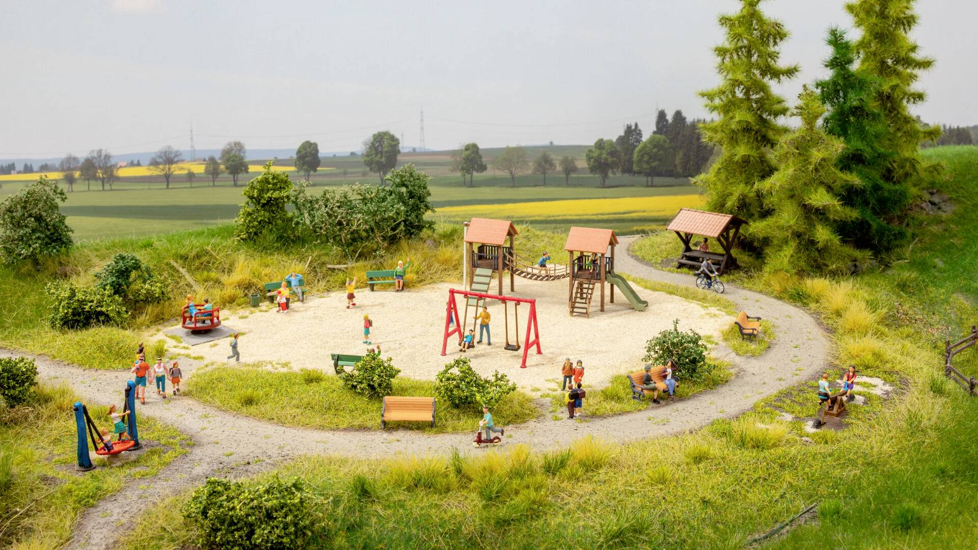 Un terrain de jeux dans un environnement rural avec des balançoires, des toboggans et des enfants qui jouent. Entouré d'arbres et de prairies.