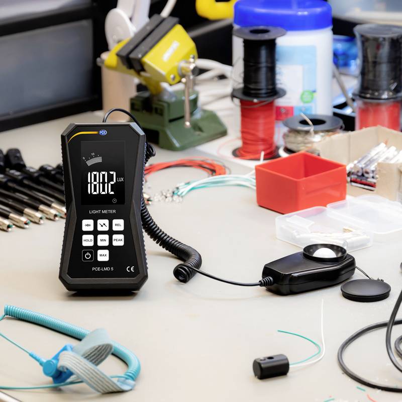 Une table montre un appareil de mesure de la lumière à 180,2 lux ainsi que des outils et des câbles pour des travaux électroniques en arrière-plan.