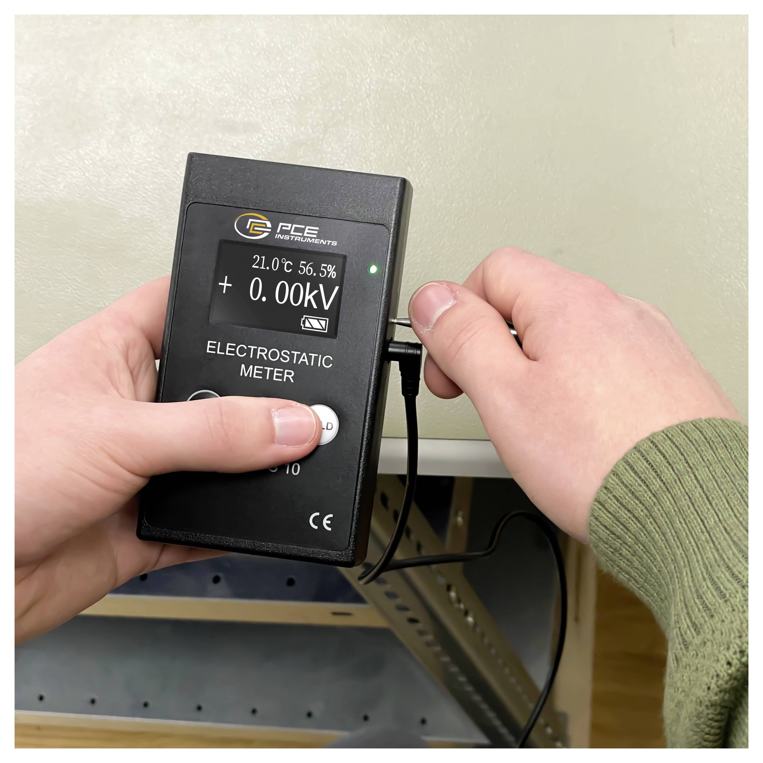 Mains utilisant un mètre électrostatique PCE pour mesurer une surface ; l'affichage indique 0,00 kV, 21,0°C et 56,5% d'humidité.