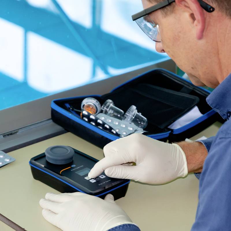 Un scientifique dans un laboratoire analyse un échantillon d'eau à l'aide d'un appareil portable, portant des gants blancs, avec du matériel de laboratoire en arrière-plan.
