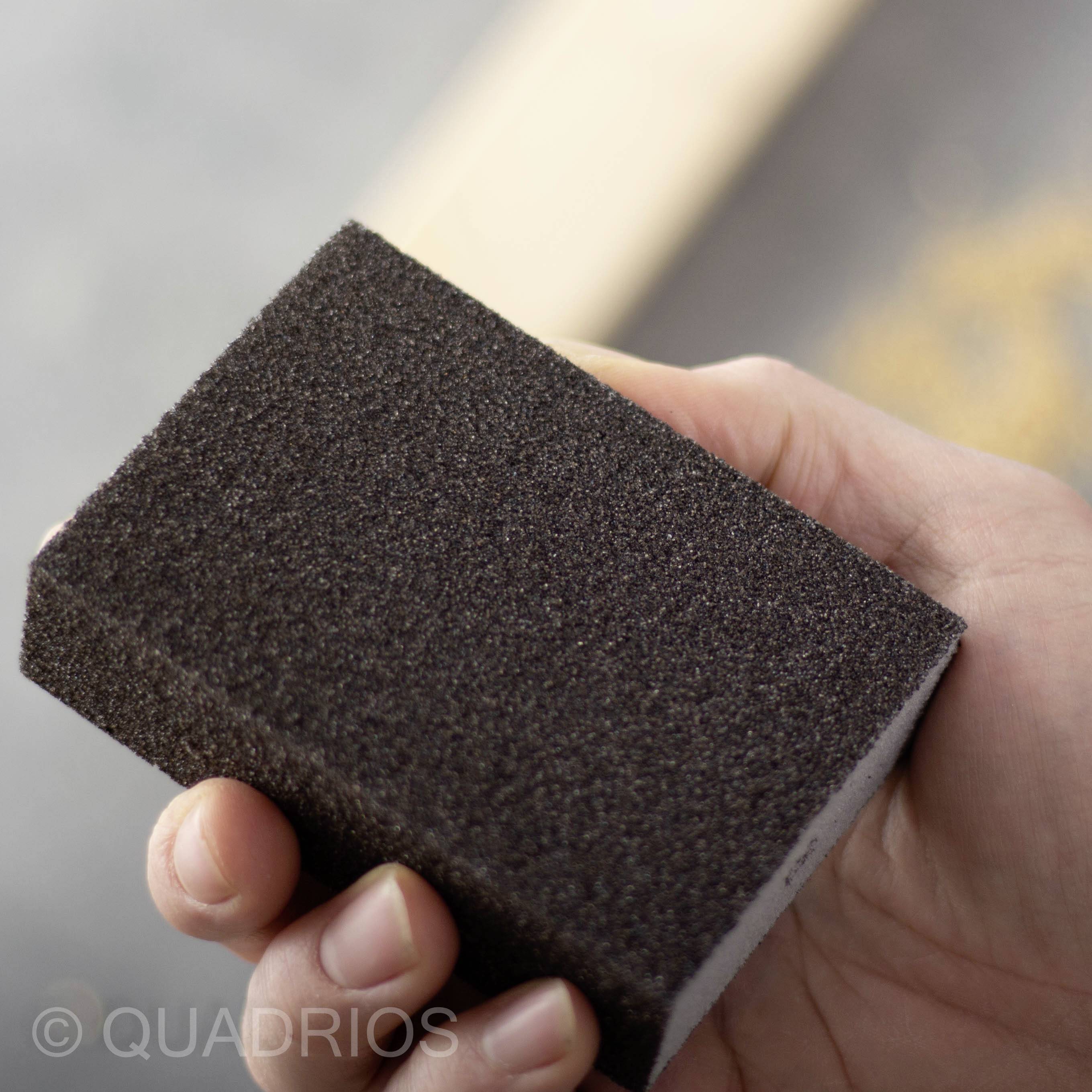 Main tenant un bloc de ponçage, prêt à être appliqué sur une surface en bois. Le bloc de ponçage est noir et rectangulaire.