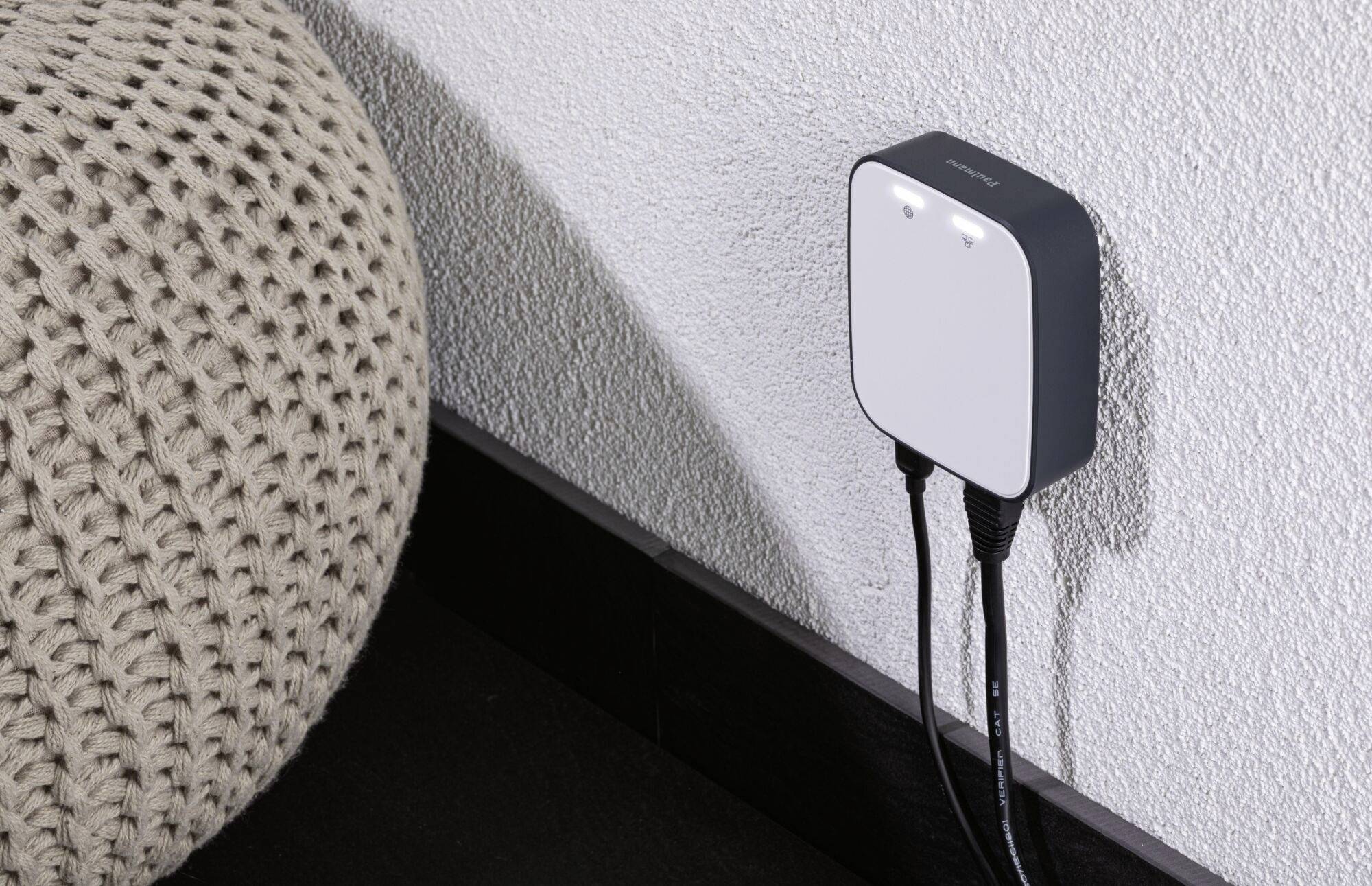 Un chargeur est branché dans une prise murale. Deux câbles y sont connectés. Un grand pouf tricoté se trouve à côté.