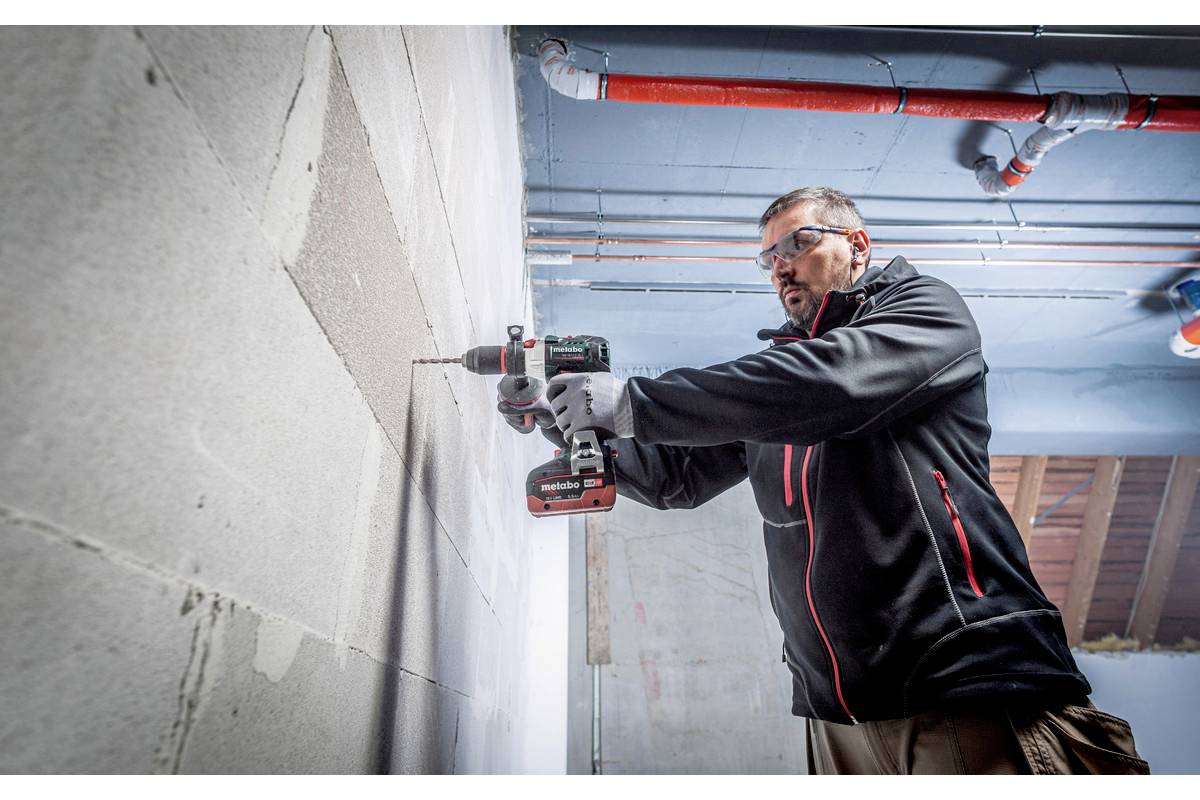Un artisan fore un trou dans un mur en béton à l'aide d'un outil électrique dans un bâtiment dont les canalisations sont apparentes au plafond.