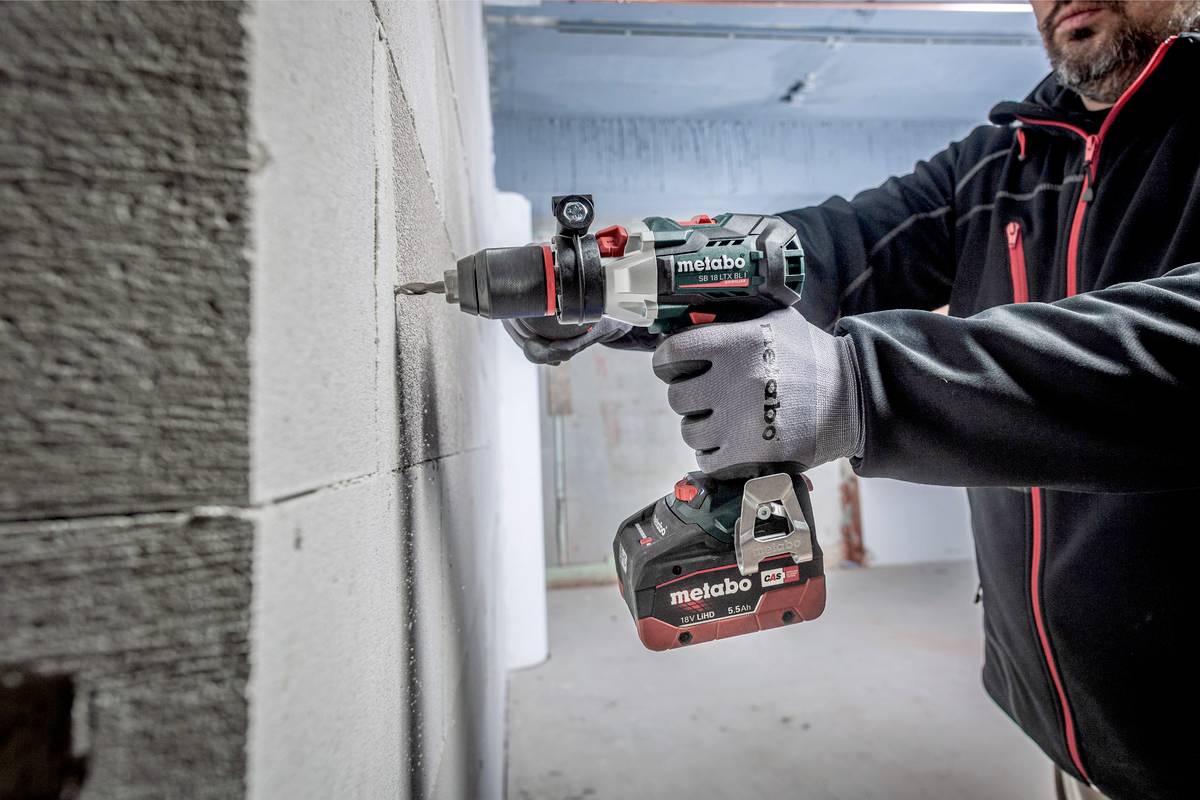 Une personne fore un mur avec une perceuse sans fil Metabo. Elle porte des vêtements de travail et des gants, la pièce semble être en état de chantier.